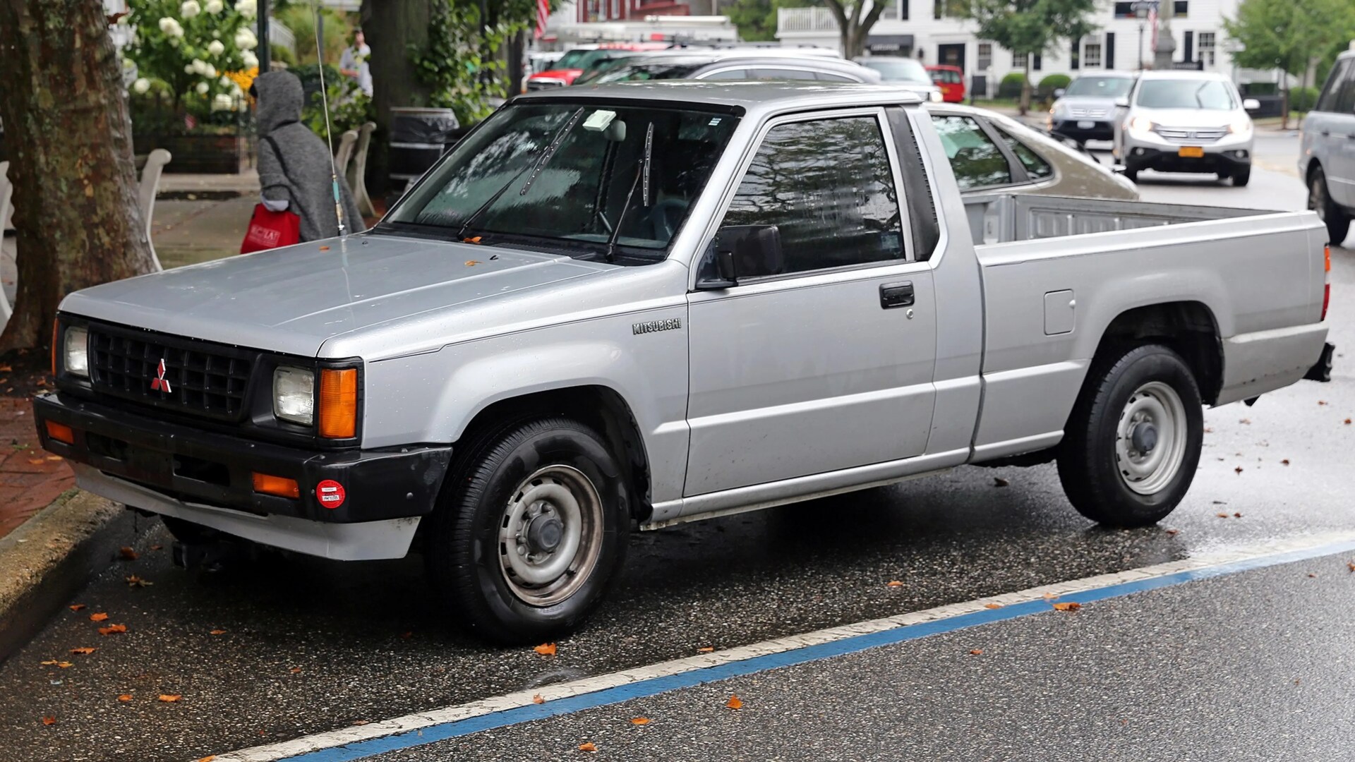 Silver Mitsubishi pickup truck parked on a wet street with a person walking nearby.