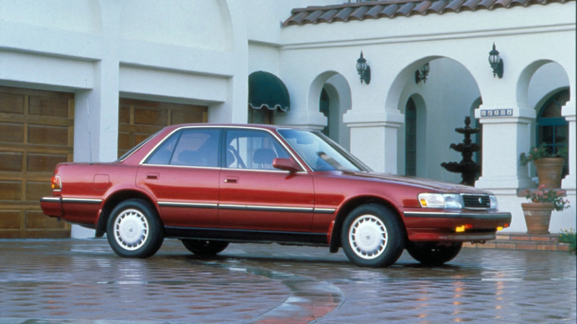 Red four-door sedan parked on a wet cobblestone driveway in front of a white building with arches and wooden garage doors.