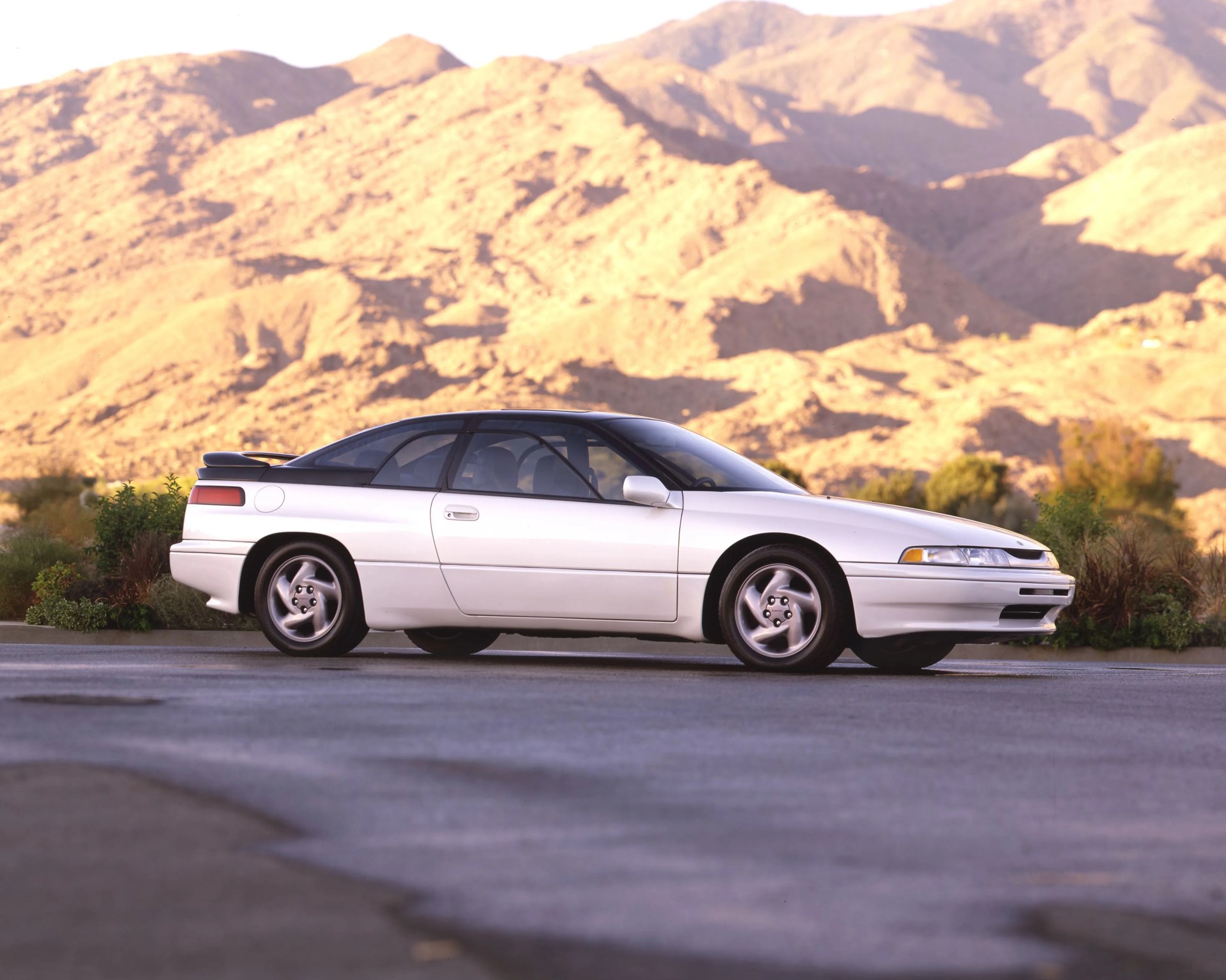 White two-door sports coupe with black roof and rear spoiler parked on asphalt with mountains in background.