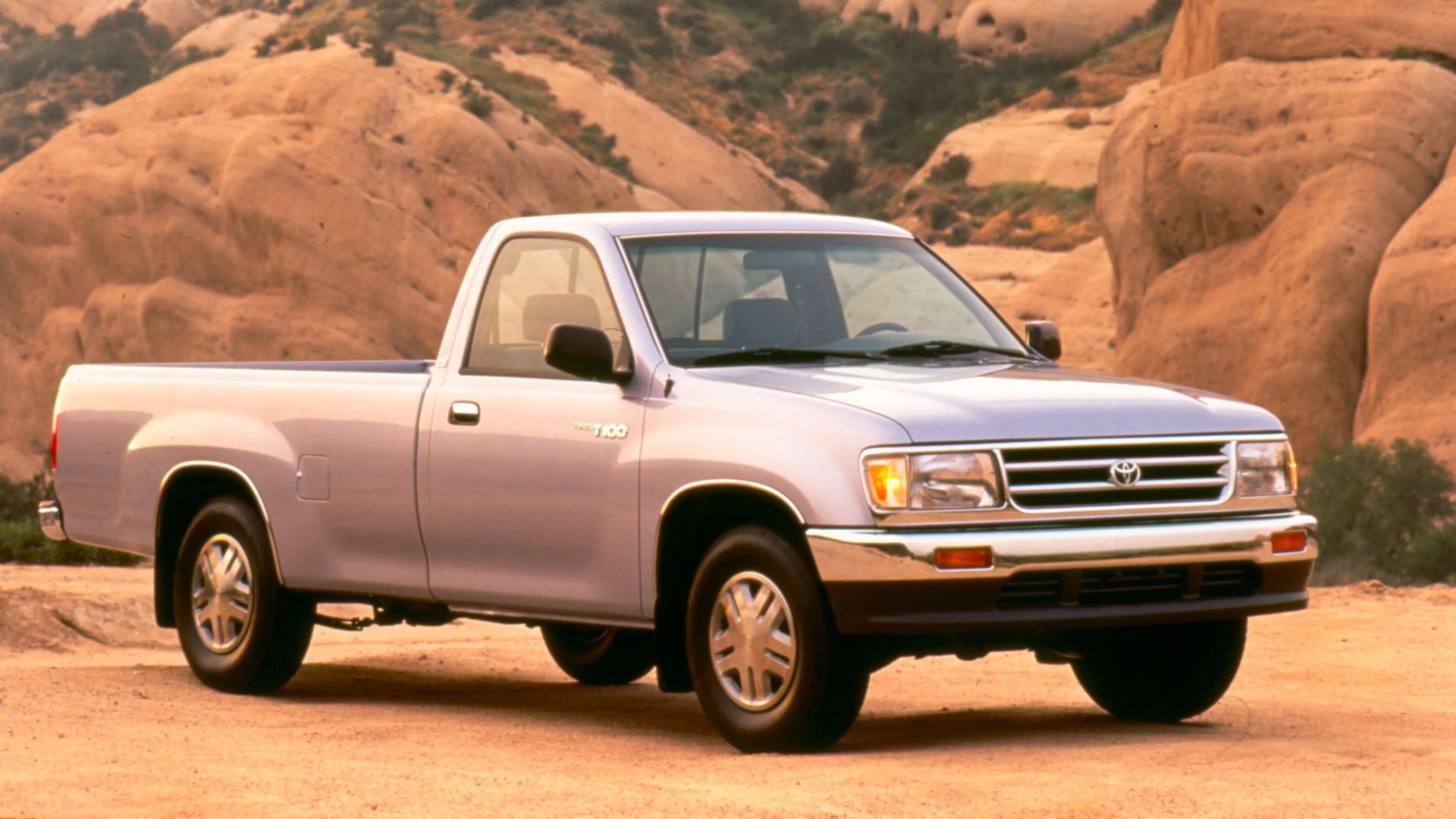 Silver Toyota T100 single-cab pickup truck parked on a dirt road with rocky hills in the background.
