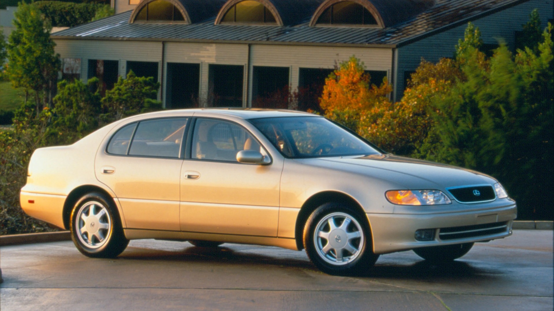 Beige Lexus sedan parked on a driveway with trees and a building in the background.