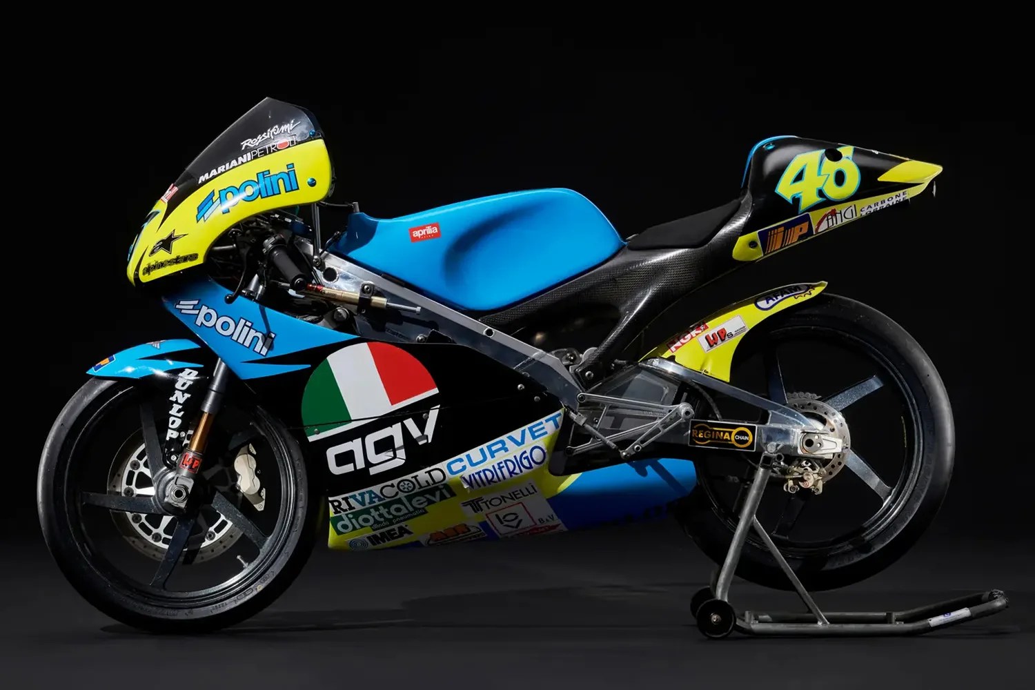Blue and yellow Aprilia racing motorcycle with sponsor logos and number 46 on a black background.