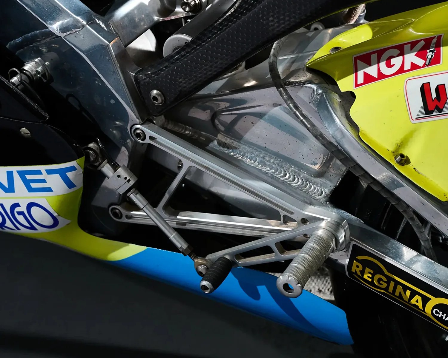 Close-up of a motorcycle's silver footpeg and gear shift lever mounted on a metallic frame with yellow and blue body panels and visible sponsor stickers.