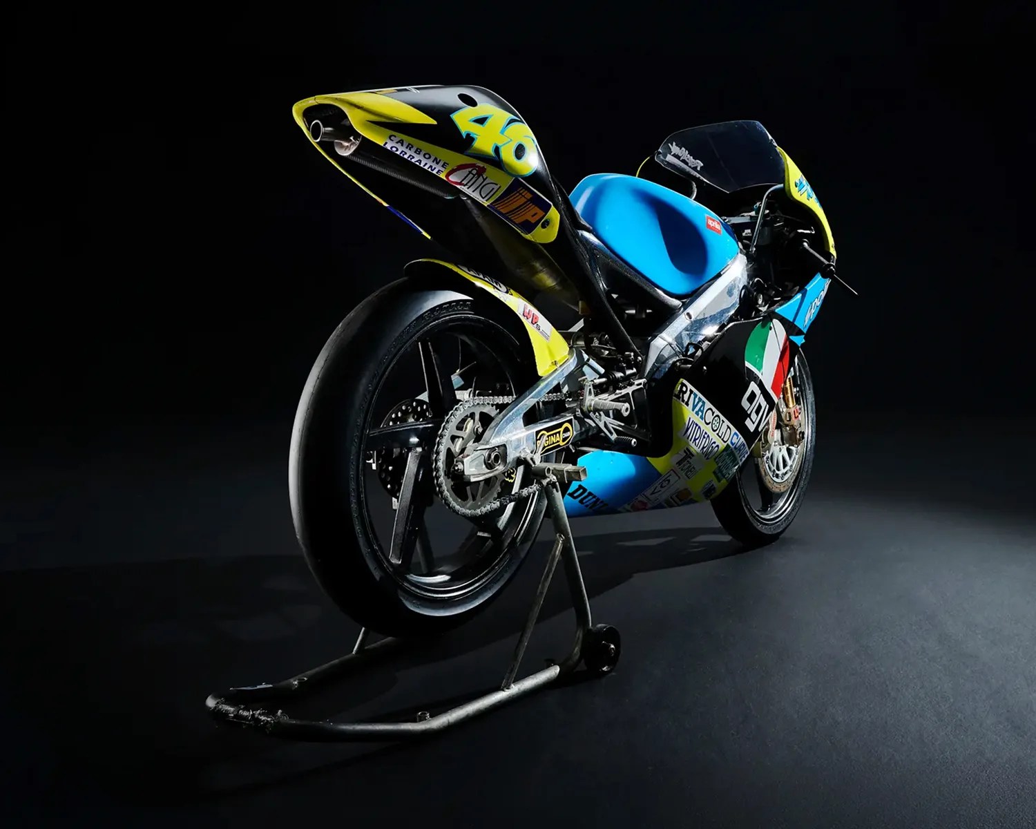 Blue and yellow racing motorcycle with sponsor decals on a black stand against a dark background.