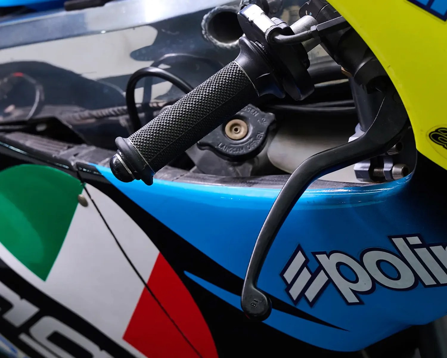 Close-up of a motorcycle handlebar grip and brake lever with blue, red, green, and white bodywork featuring the "polini" logo.
