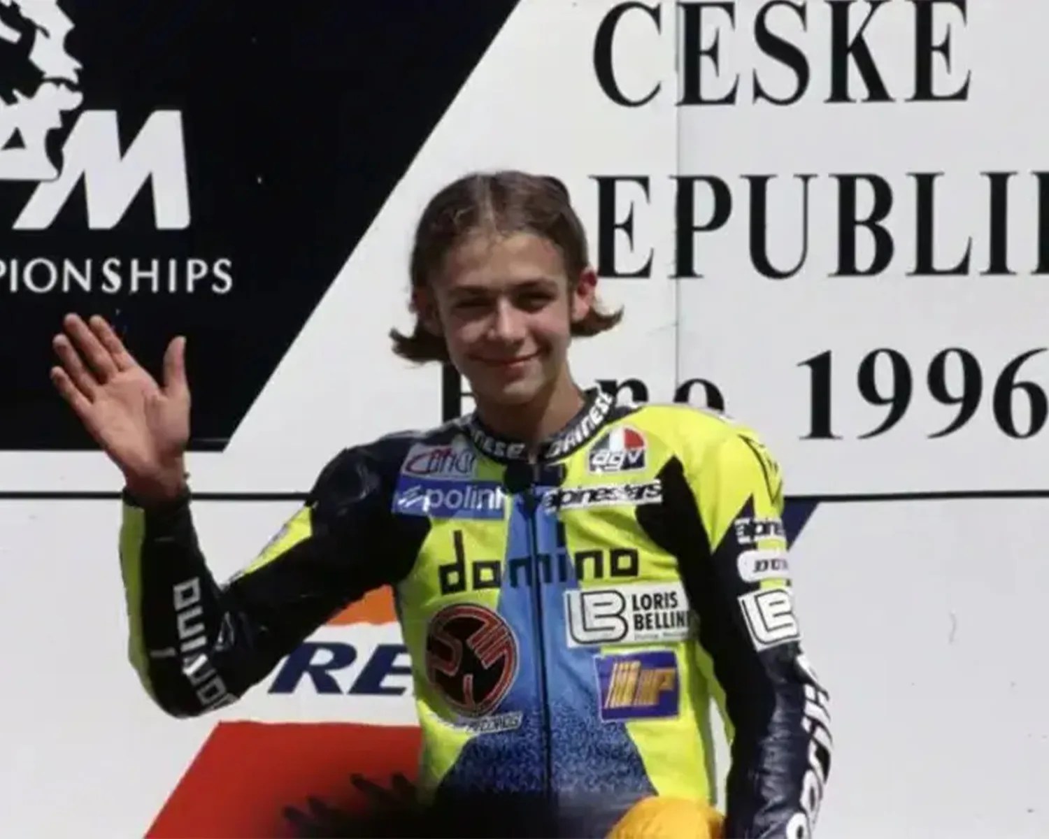 Young motorcyclist in a yellow and blue racing suit waving with a backdrop showing "CESKE REPUBLIK" and "1996".