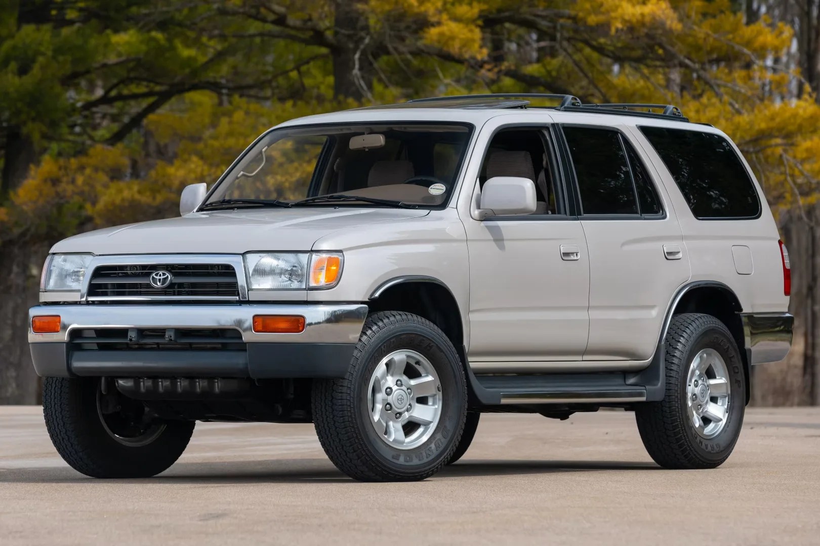 White Toyota 4Runner SUV with chrome bumper and roof rack parked on pavement with autumn trees in background.