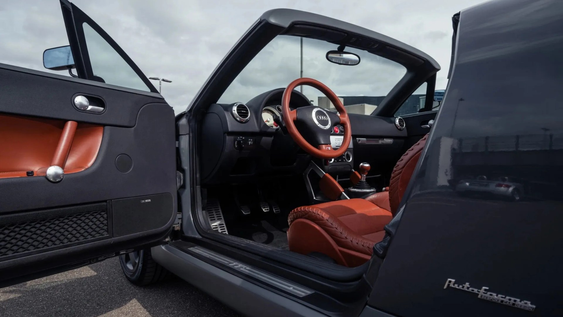 Open black Audi convertible car door showing brown leather interior and steering wheel with Audi logo.