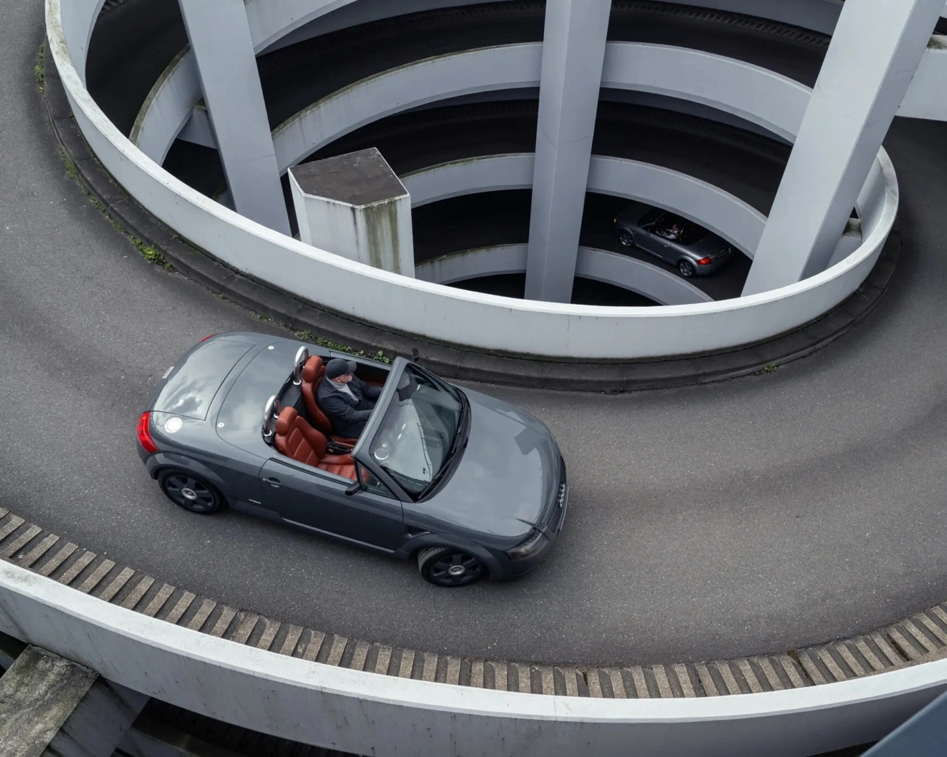 Gray convertible car with brown leather seats driving on a spiral parking ramp.