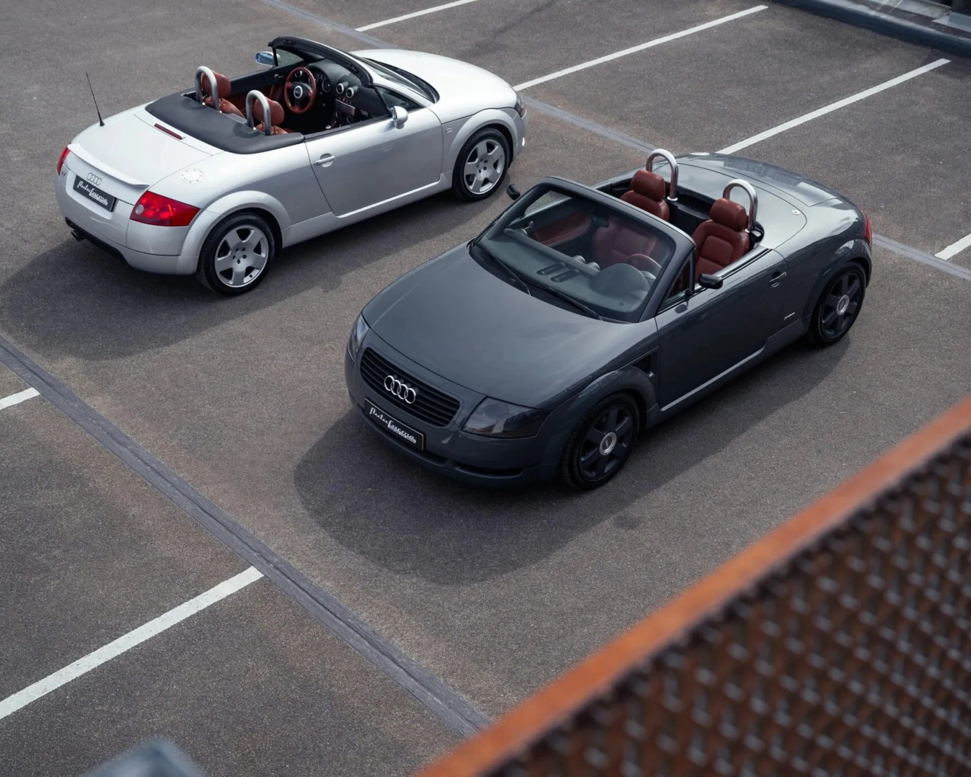 Two Audi TT convertible cars, one silver and one dark gray, parked in an empty lot with red leather interiors.