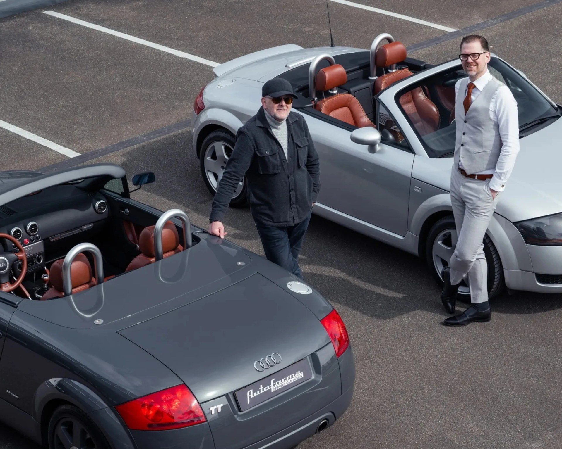 Two men standing near two convertible Audi TT cars, one silver and one dark gray, in a parking lot.