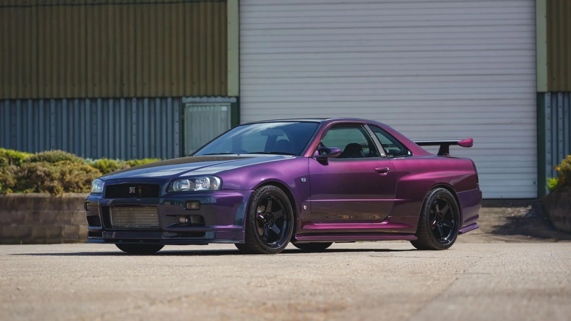 Purple Nissan GT-R R34 with black wheels parked in front of a corrugated metal building.