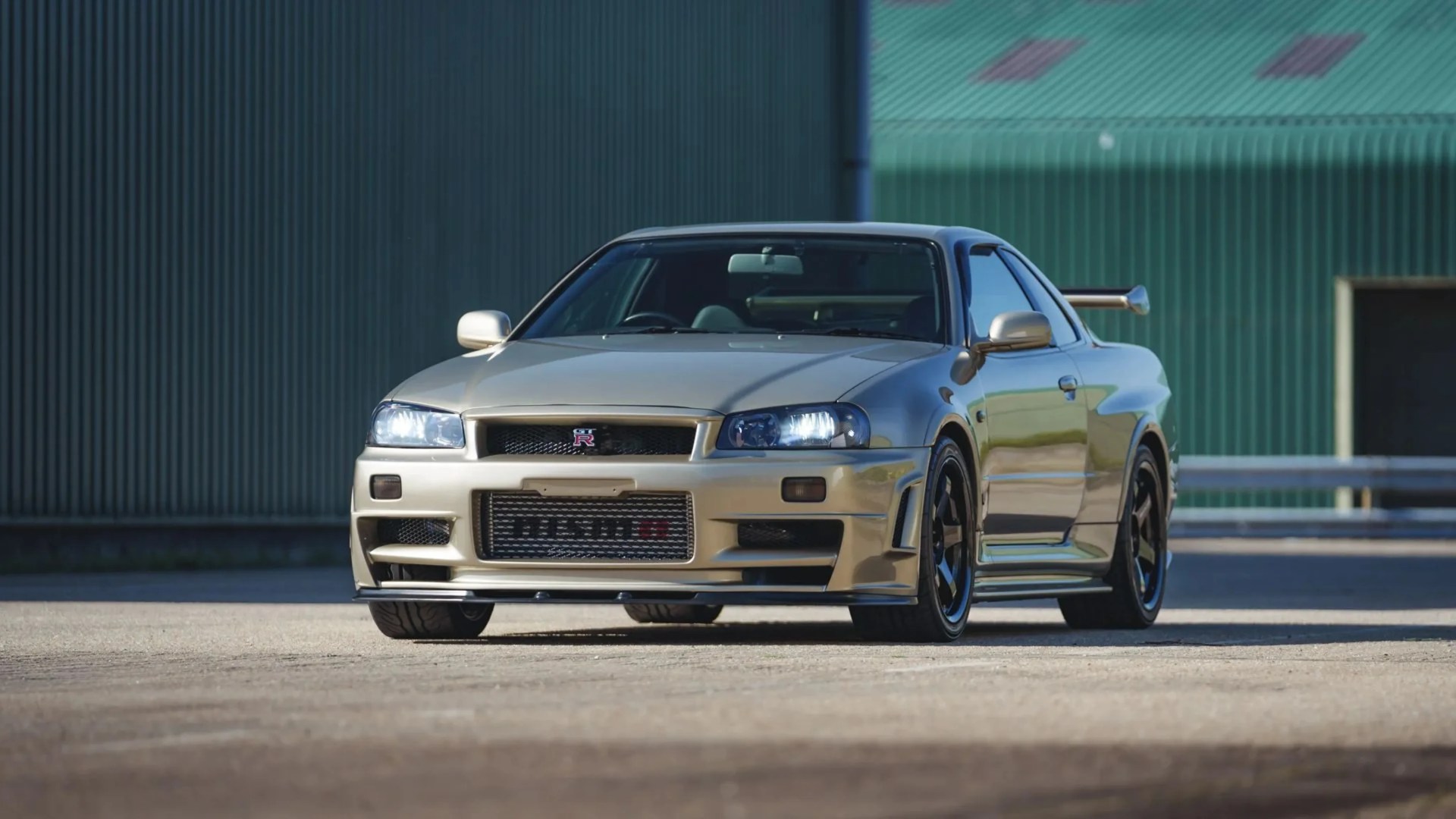 Champagne gold Nissan Skyline GT-R R34 with black wheels parked on pavement in front of green industrial building.