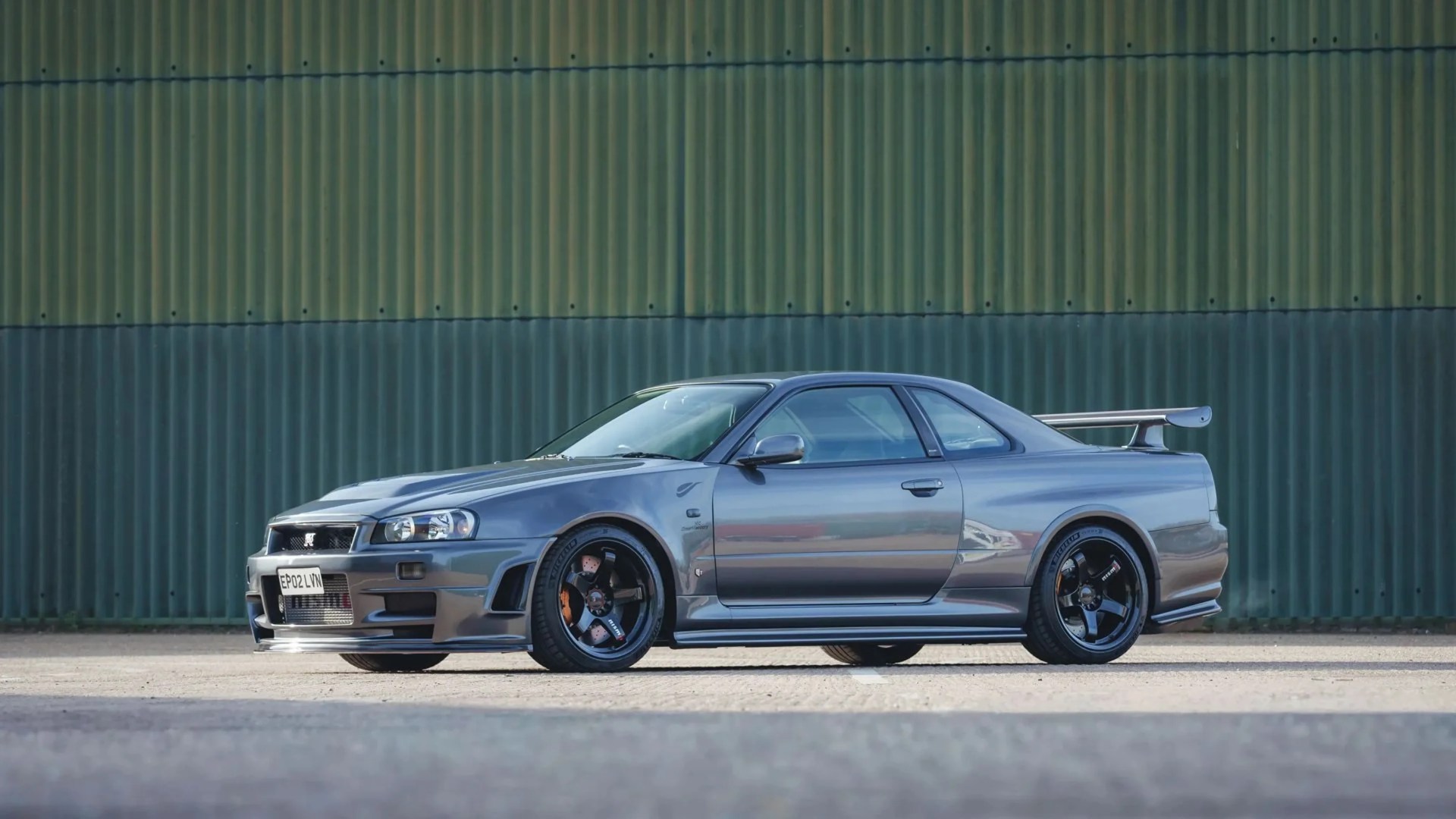Gray Nissan Skyline GT-R R34 coupe with black wheels and rear spoiler parked in front of a green corrugated metal wall.
