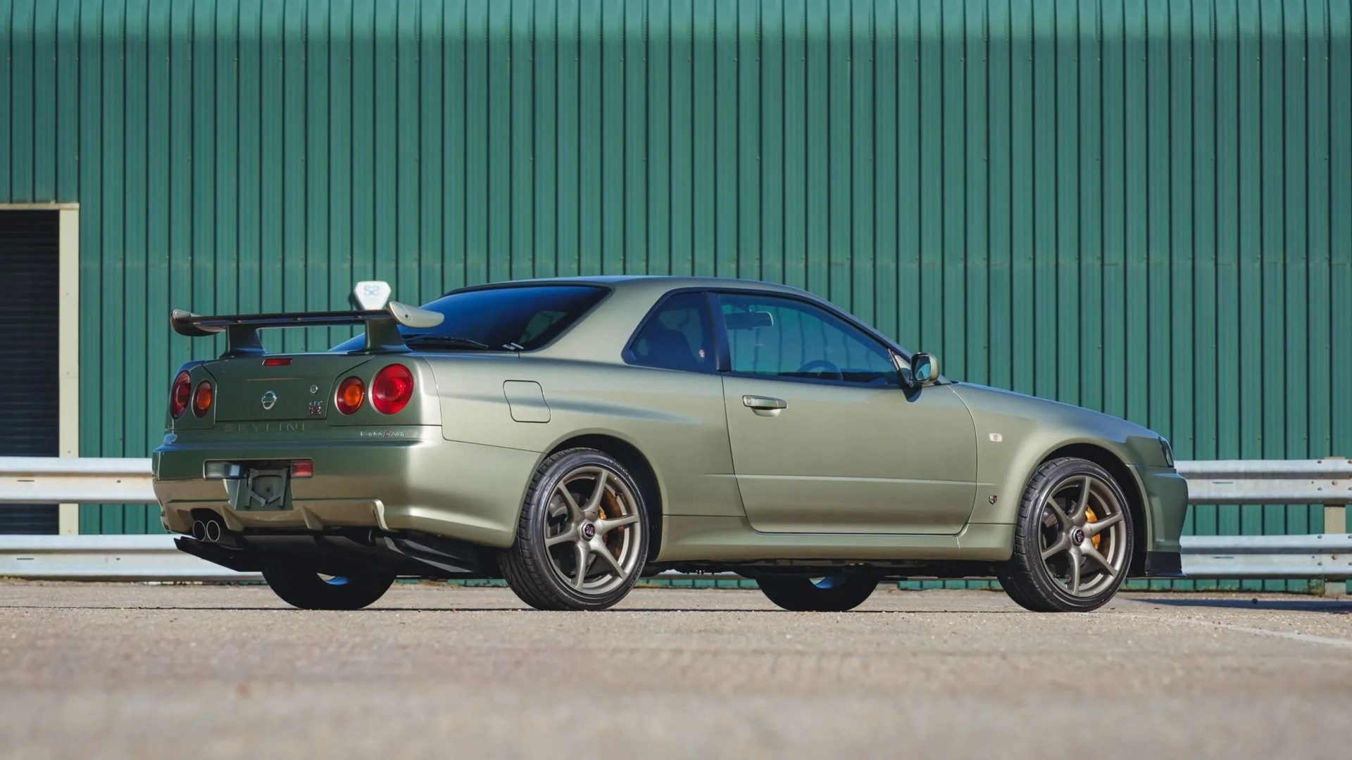 Metallic green Nissan Skyline GT-R R34 coupe with rear spoiler parked on pavement in front of green corrugated metal wall.