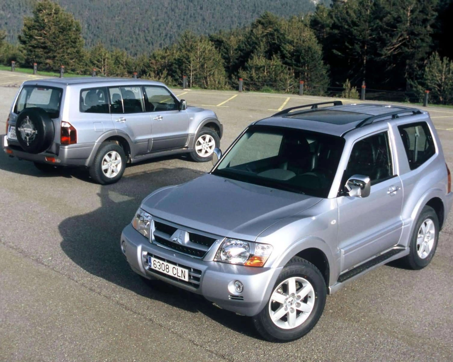 Two silver Mitsubishi Pajero SUVs parked on an asphalt lot with trees in the background.