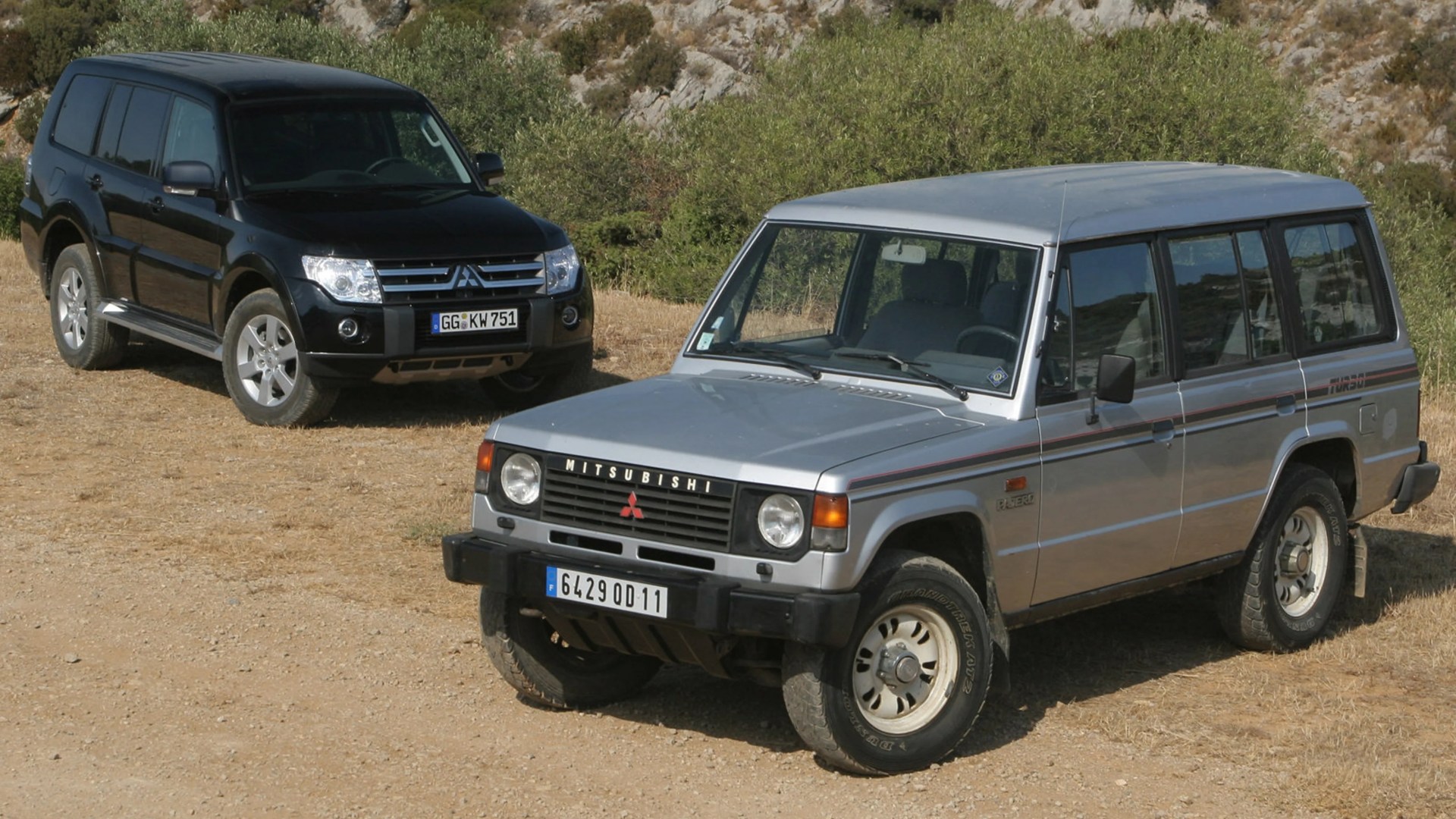 Two Mitsubishi Pajero SUVs, one silver older model in the foreground and one black newer model in the background, parked on dry dirt with greenery behind.