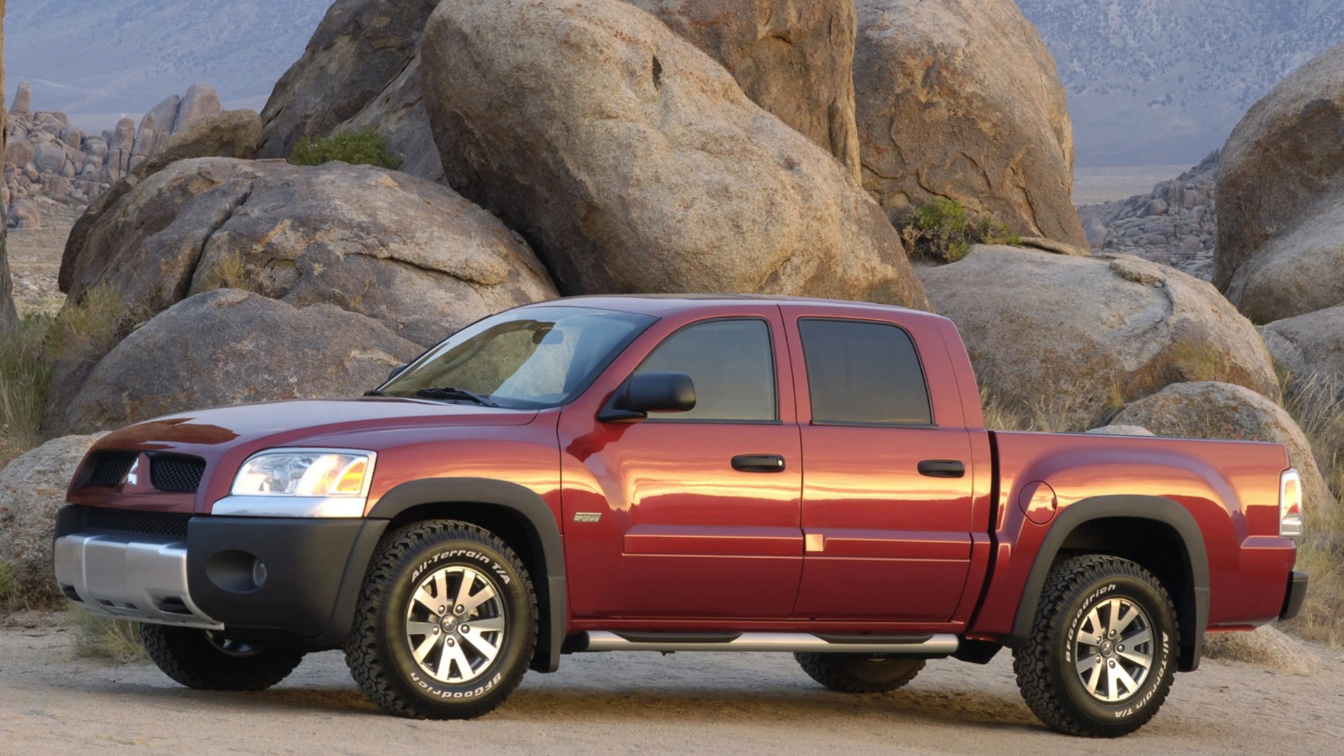 Red Mitsubishi pickup truck parked on a dirt road with large rocks in the background.