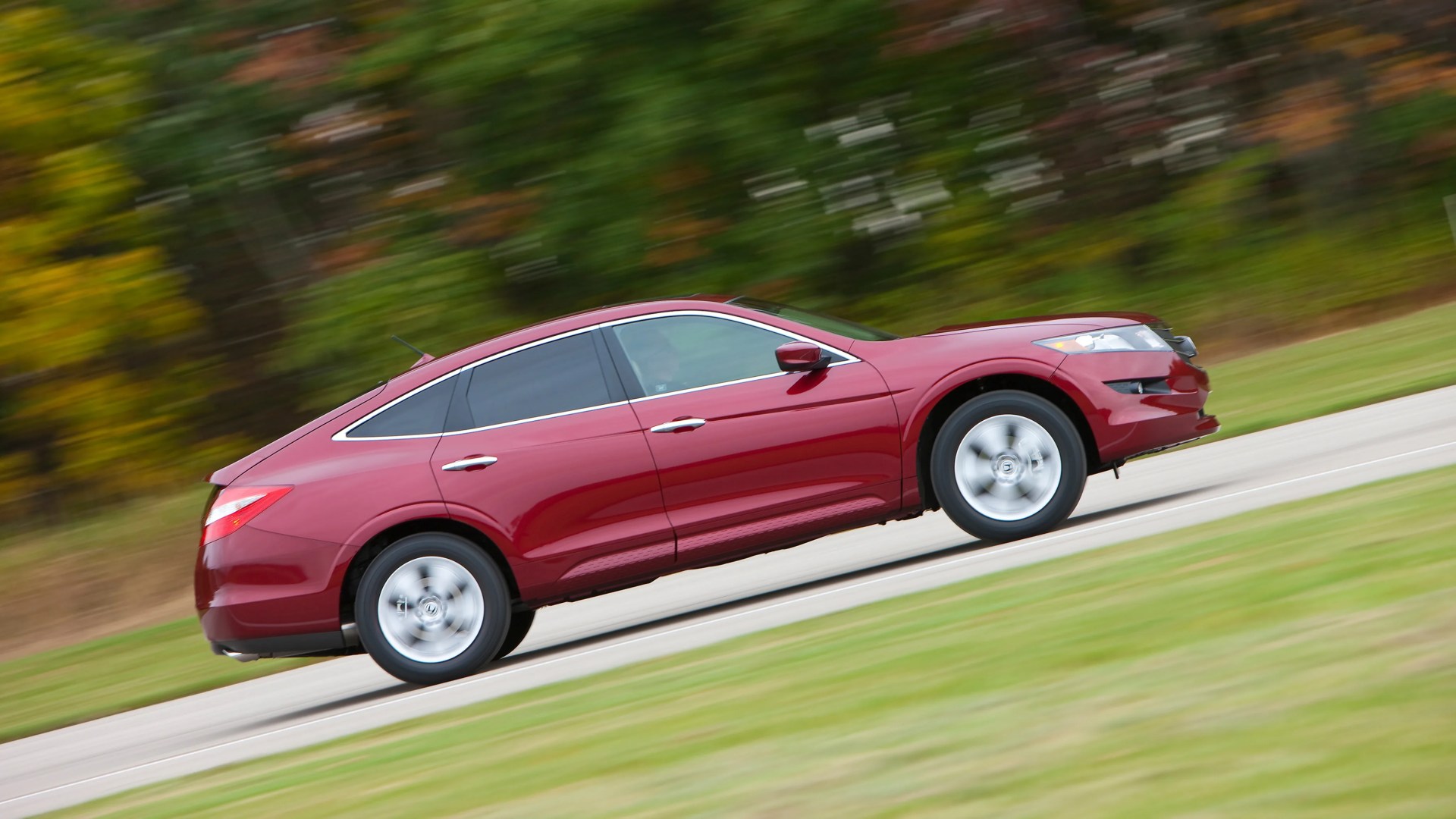 Red four-door sedan driving on a road with blurred green and yellow foliage in the background.