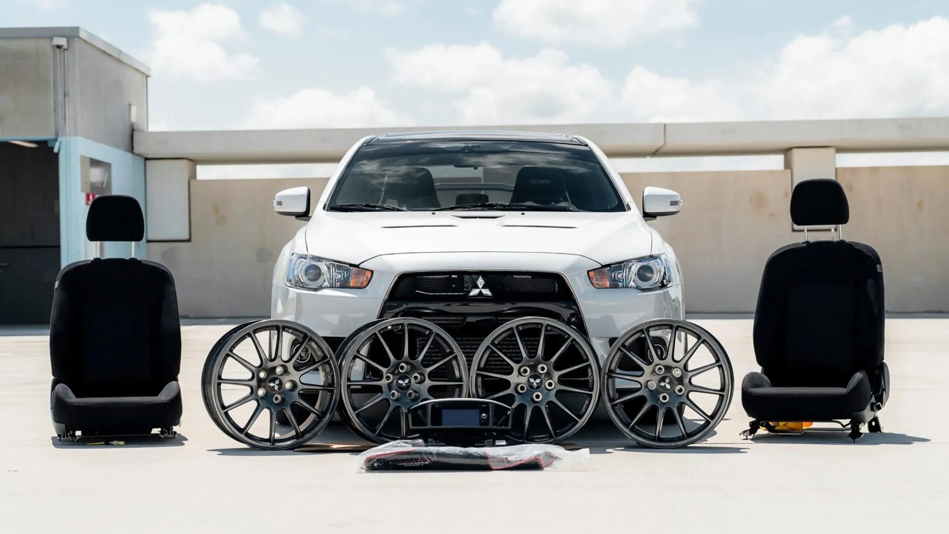 White Mitsubishi SUV with four black alloy wheels and two black car seats arranged in front on a concrete surface.