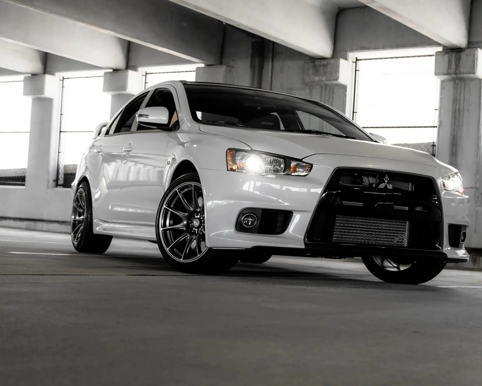 White Mitsubishi Lancer Evolution with black grille and alloy wheels in a concrete parking garage.