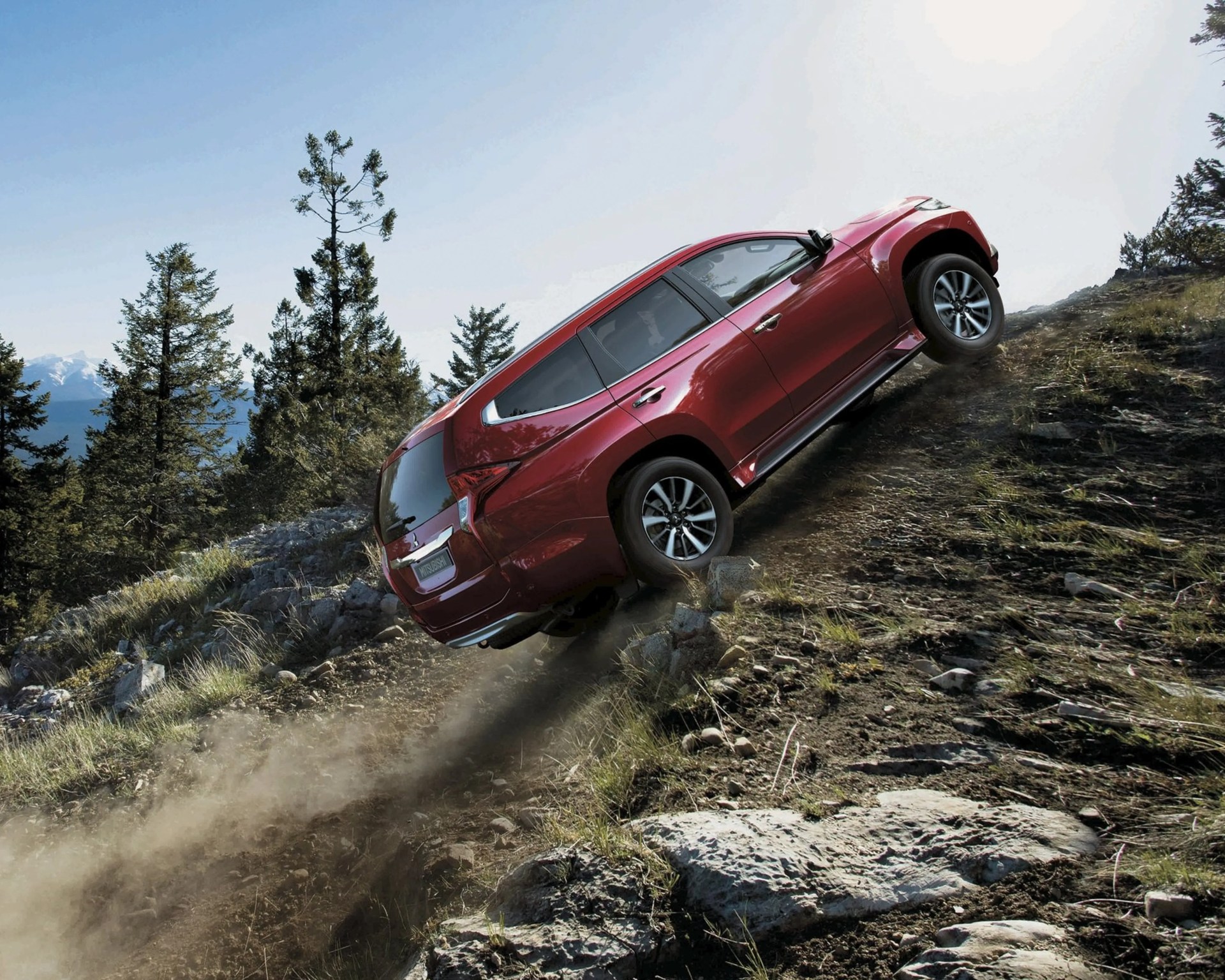 Red Mitsubishi SUV climbing a steep rocky dirt hill with trees and mountains in the background.