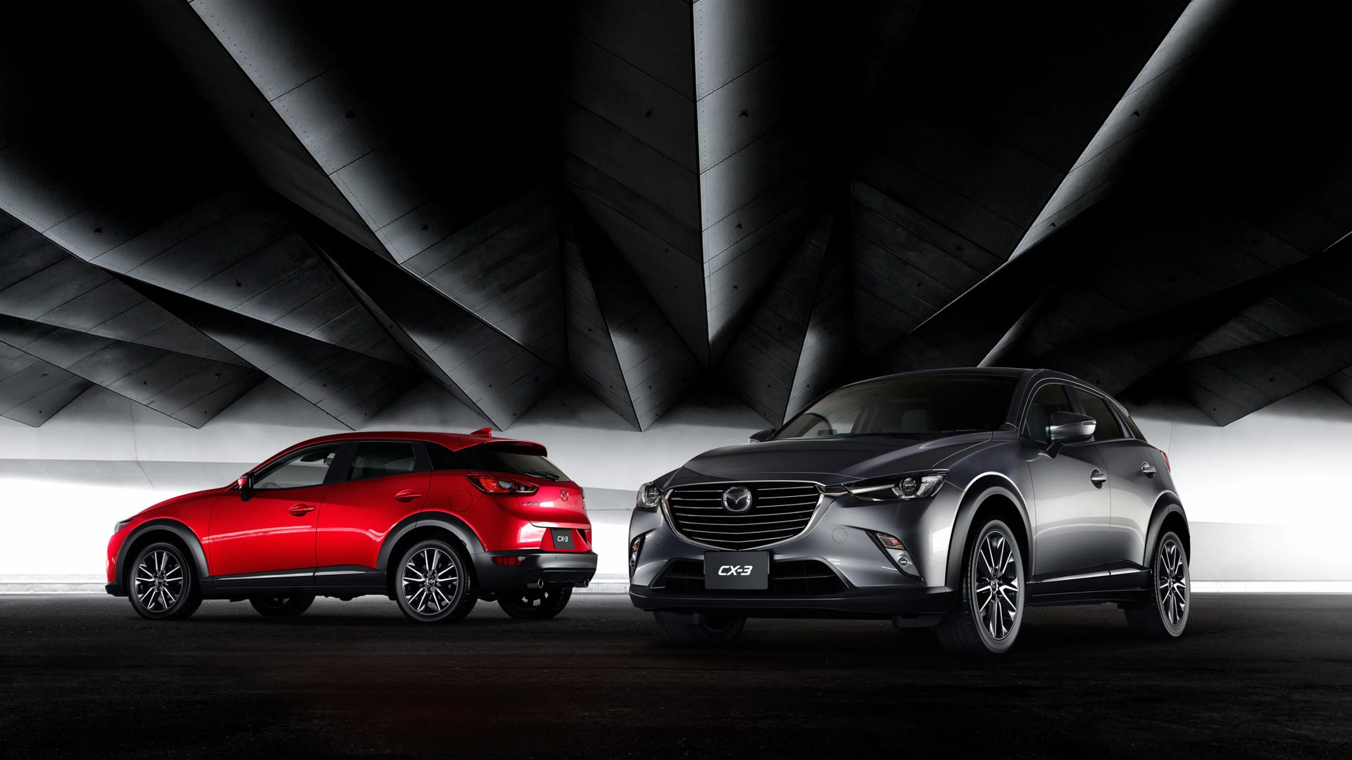Two Mazda CX-3 SUVs, one red and one gray, parked under a modern, angular concrete ceiling.