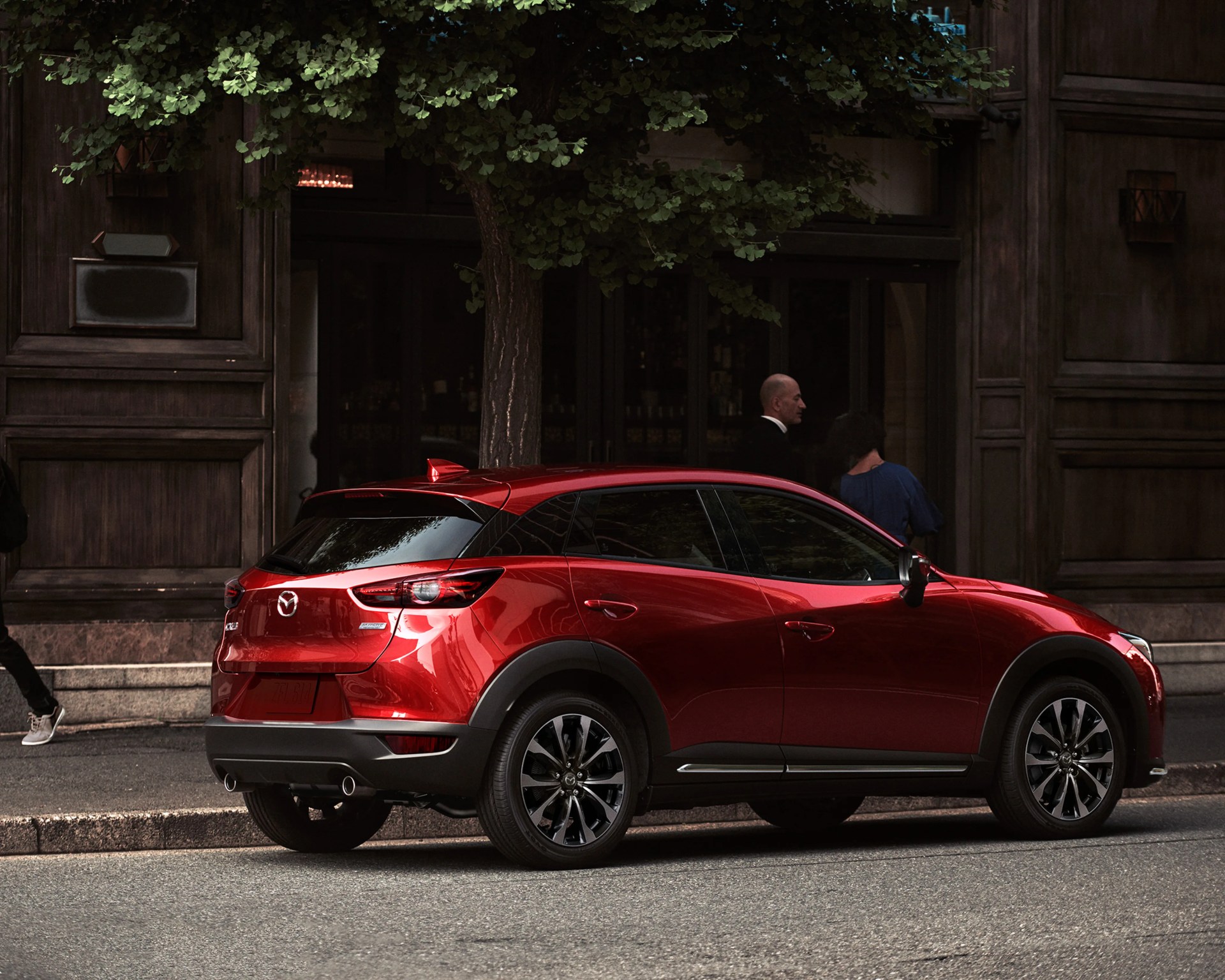 Red Mazda CX-30 SUV parked on a street with dark wood building and green tree in the background.