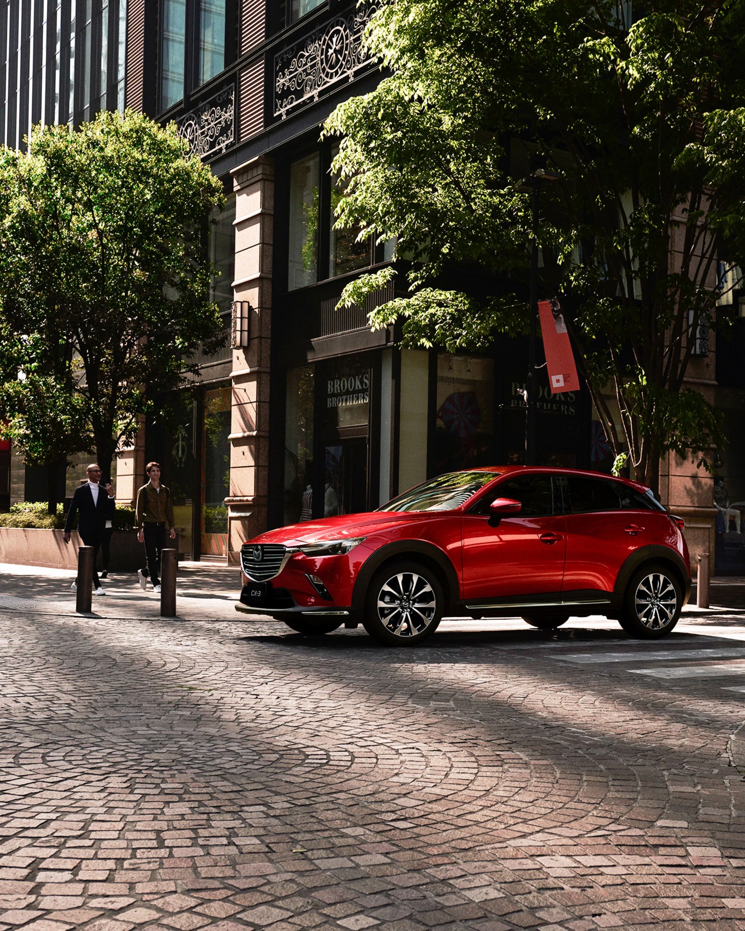 Red Mazda CX-3 compact SUV parked on a cobblestone street near a building with trees and two people walking nearby.