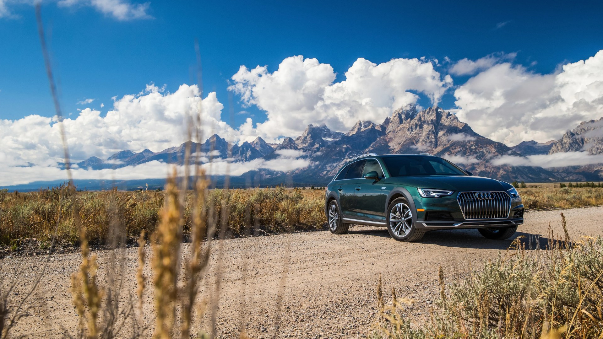 Dark green Audi SUV parked on a dirt road with mountains and blue sky in the background.