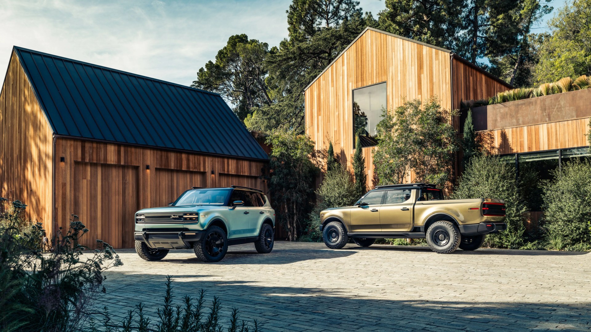 Two modern SUVs, one light blue and one gold, parked on a stone driveway in front of a wooden house with black metal roof.