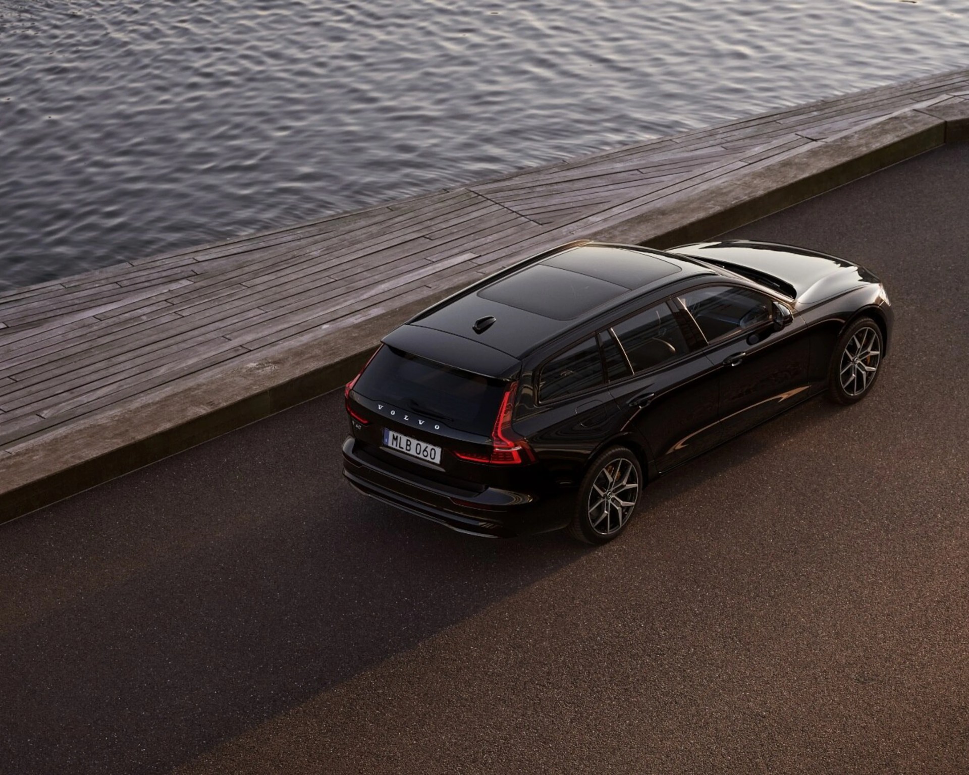 Black Volvo V60 station wagon parked on asphalt near a wooden dock by the water.