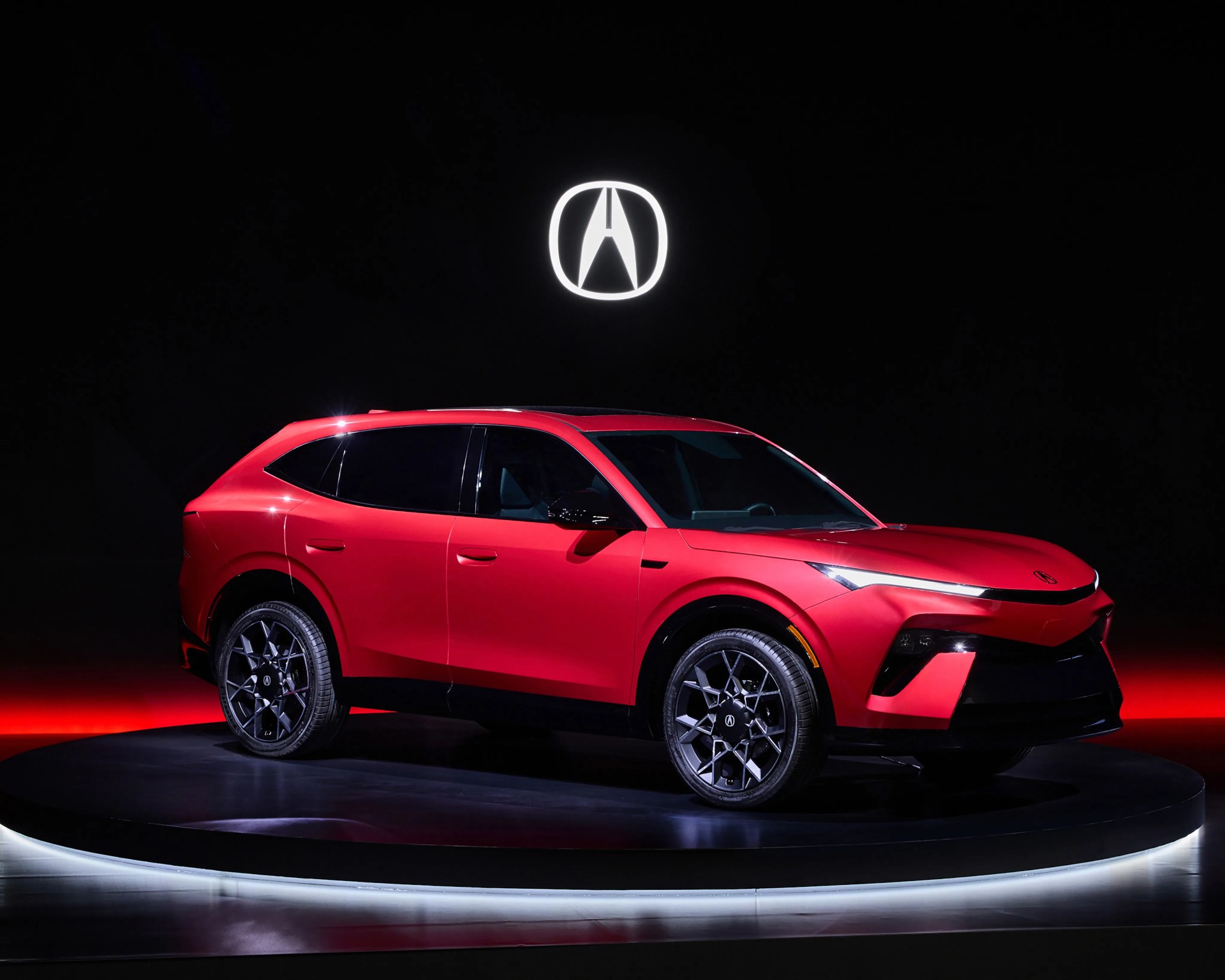 Red Acura SUV with black wheels on a circular platform under a glowing Acura logo on a black background.