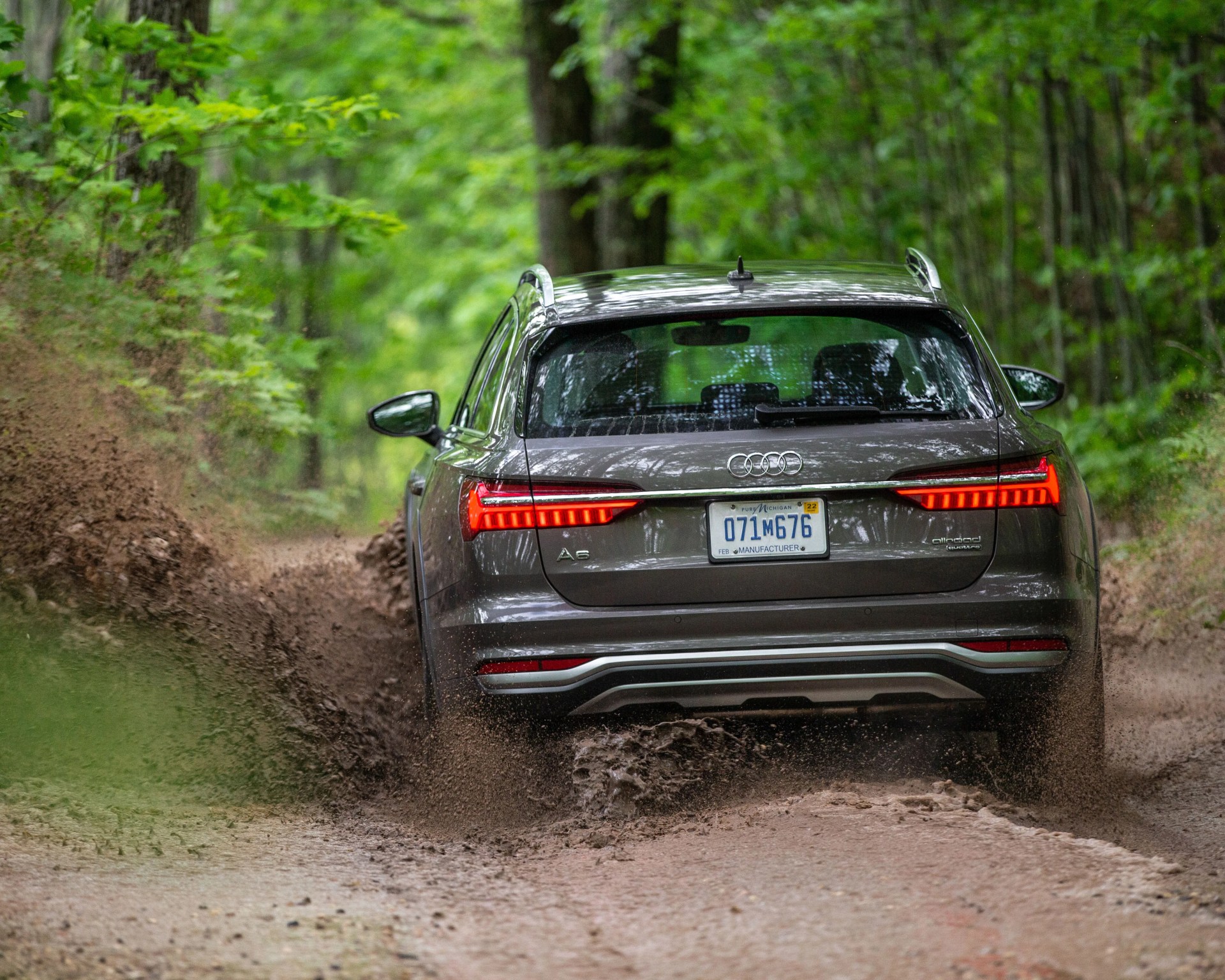 Gray Audi A6 allroad quattro driving on a muddy forest trail with mud splashing behind.