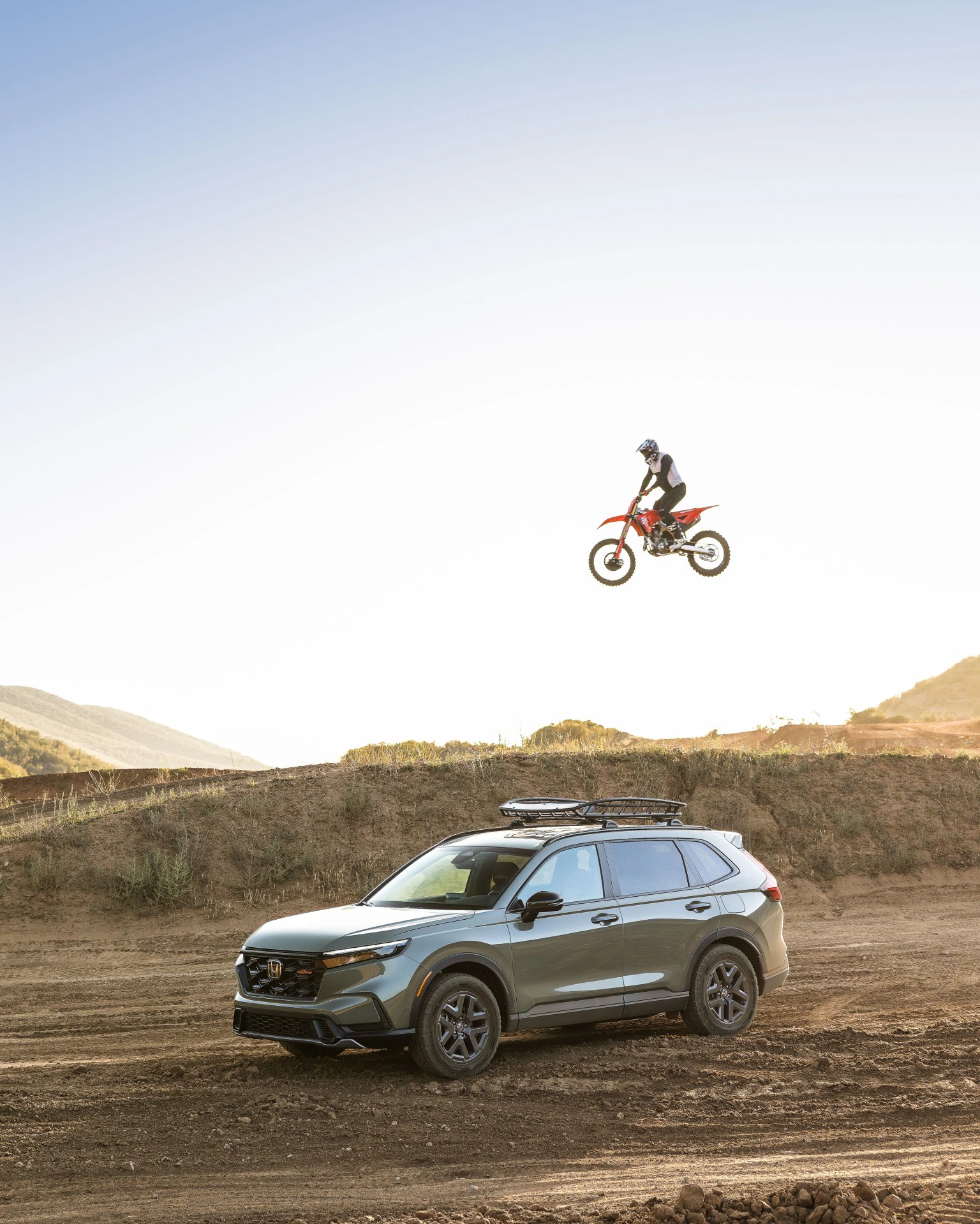 Gray Honda SUV parked on dirt terrain with a motocross rider jumping on a red dirt bike in the background.