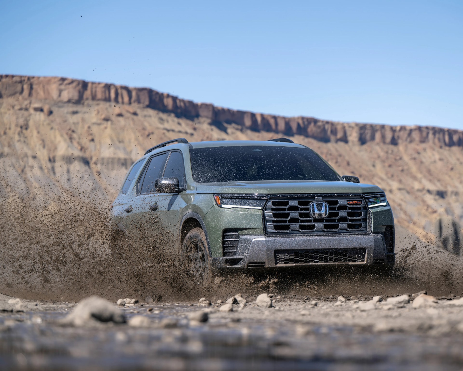 Green Honda SUV driving through muddy terrain with dirt splashing around, rocky cliffs in background.