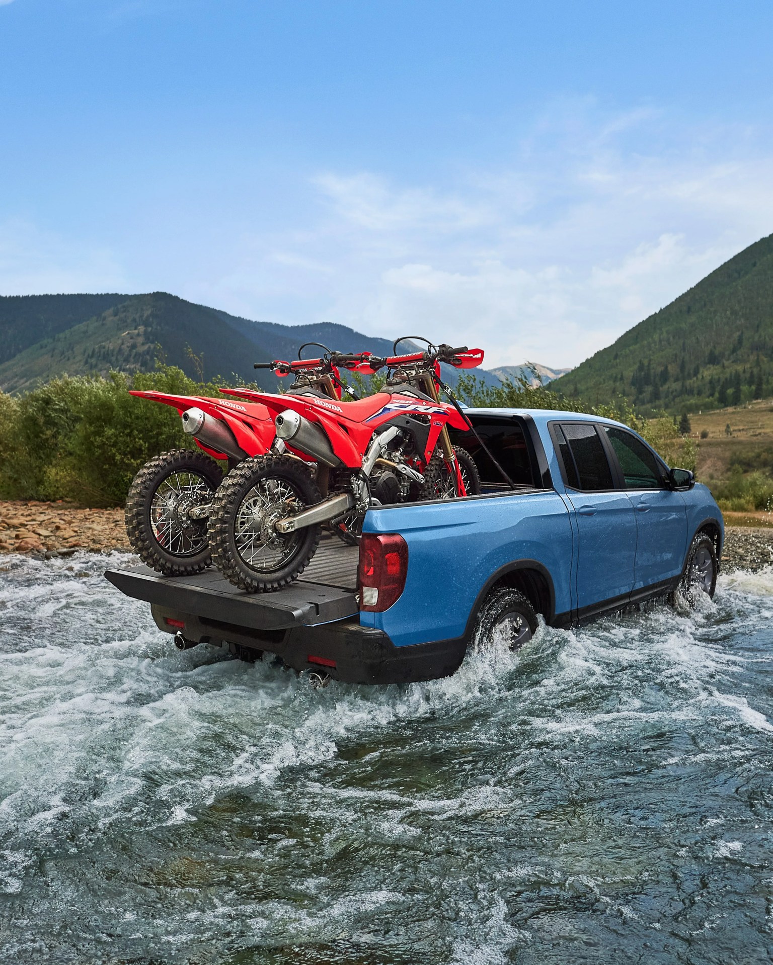 Blue pickup truck driving through water with two red Honda dirt bikes in the bed, mountains in background.