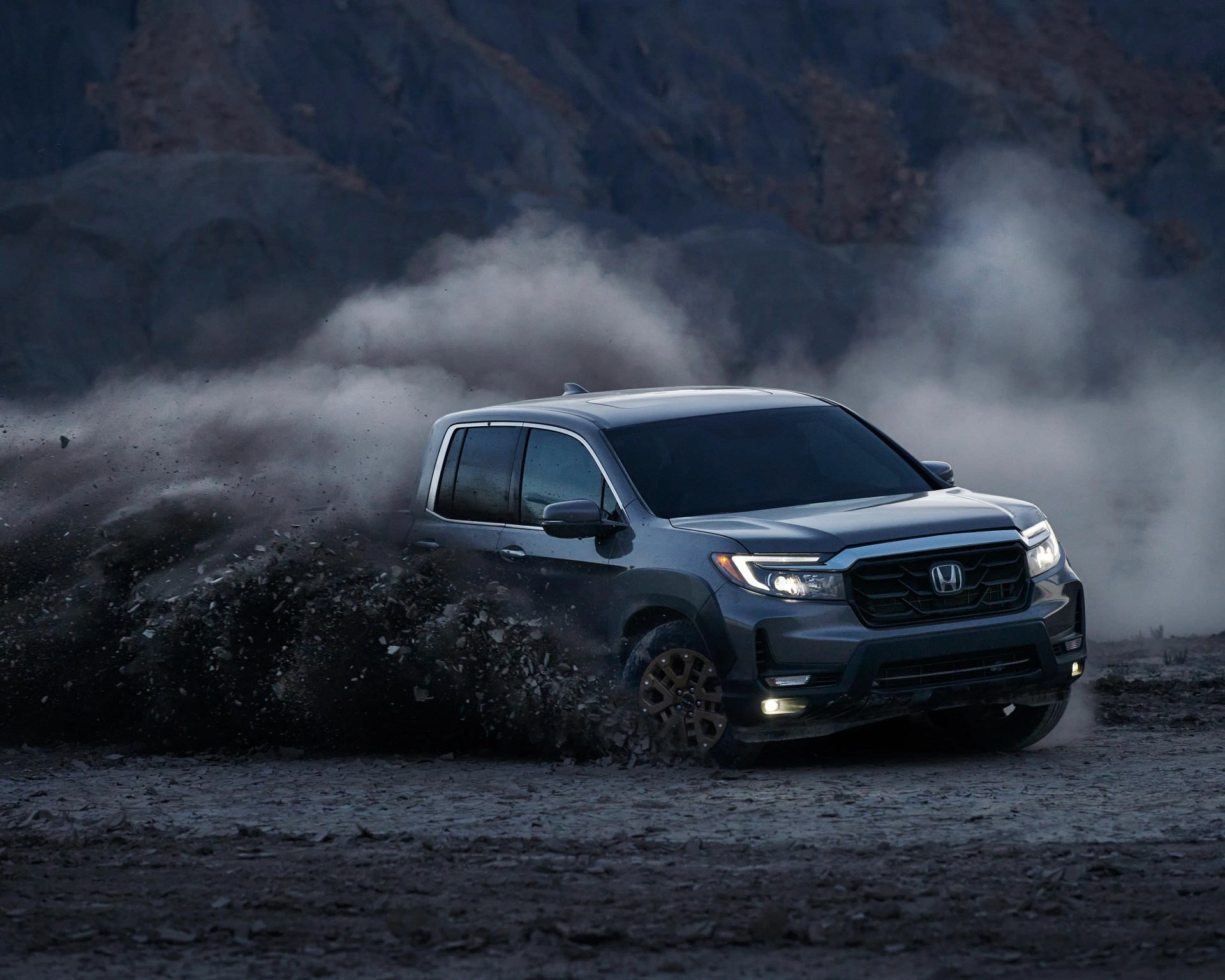 Gray Honda pickup truck kicking up dust and rocks on a rugged dirt terrain with mountains in the background.