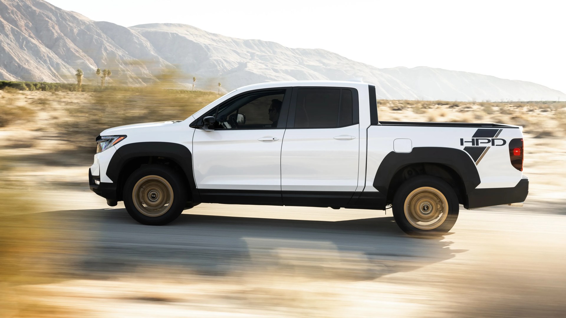 White pickup truck with black trim and bronze wheels driving in a desert landscape with mountains in the background.