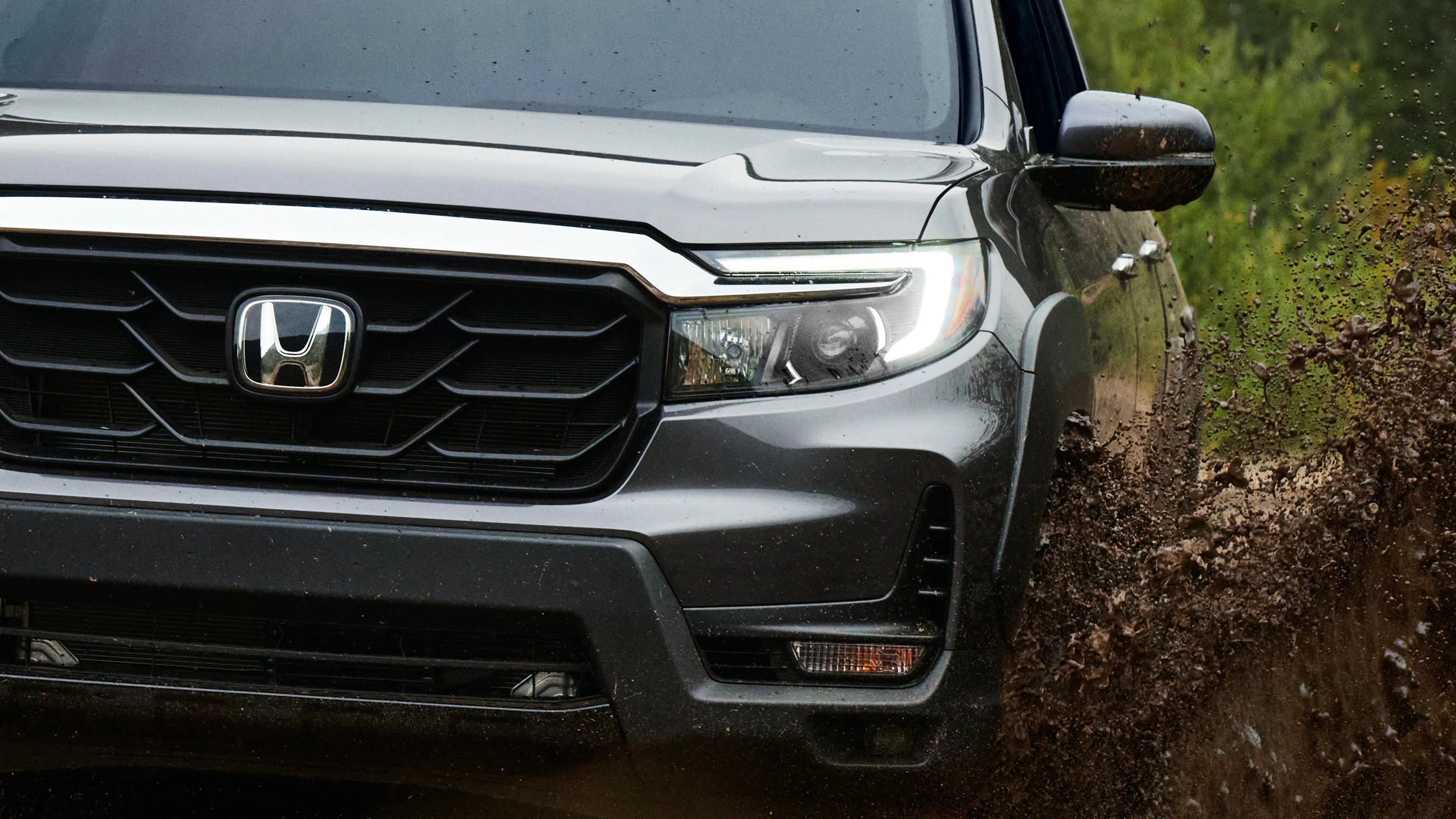 Front view of a gray Honda SUV splashing mud on a dirt road with LED headlights on.