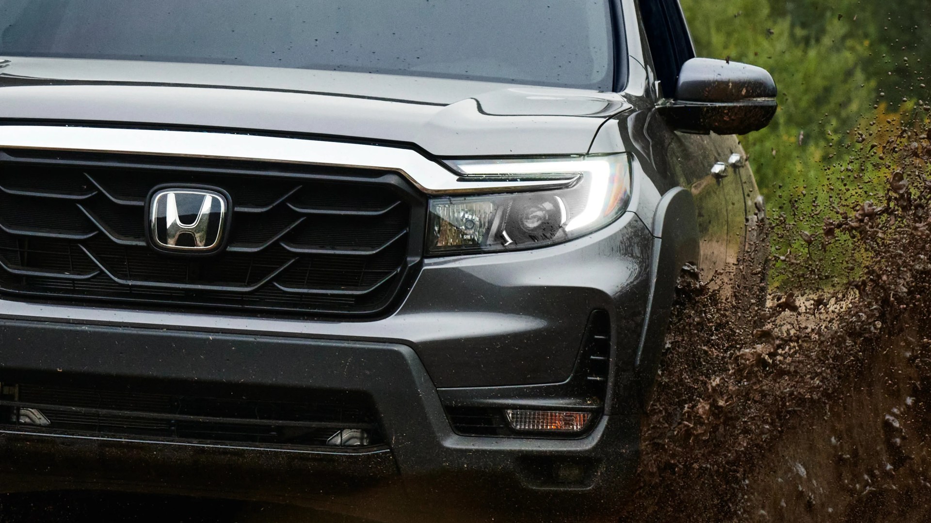Front view of a gray Honda SUV splashing mud on a dirt road with LED headlights on.