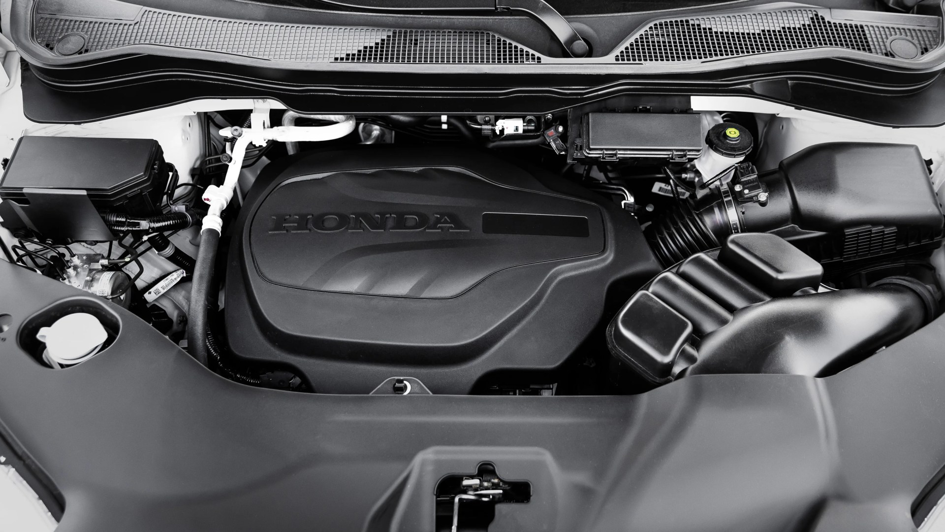 Black plastic engine cover with "HONDA" embossed, surrounded by various engine components in a car engine bay.