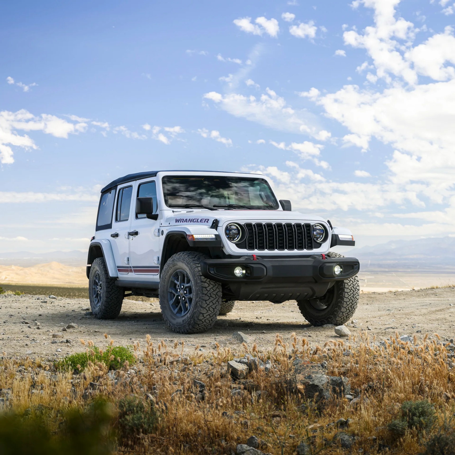 White Jeep Wrangler with black soft top parked on rocky terrain under a partly cloudy blue sky.
