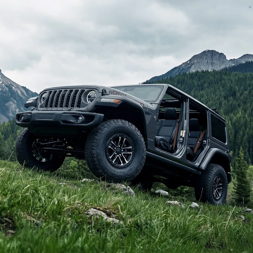 Black Jeep Wrangler with open doors parked on grassy hill with forest and mountains in the background.