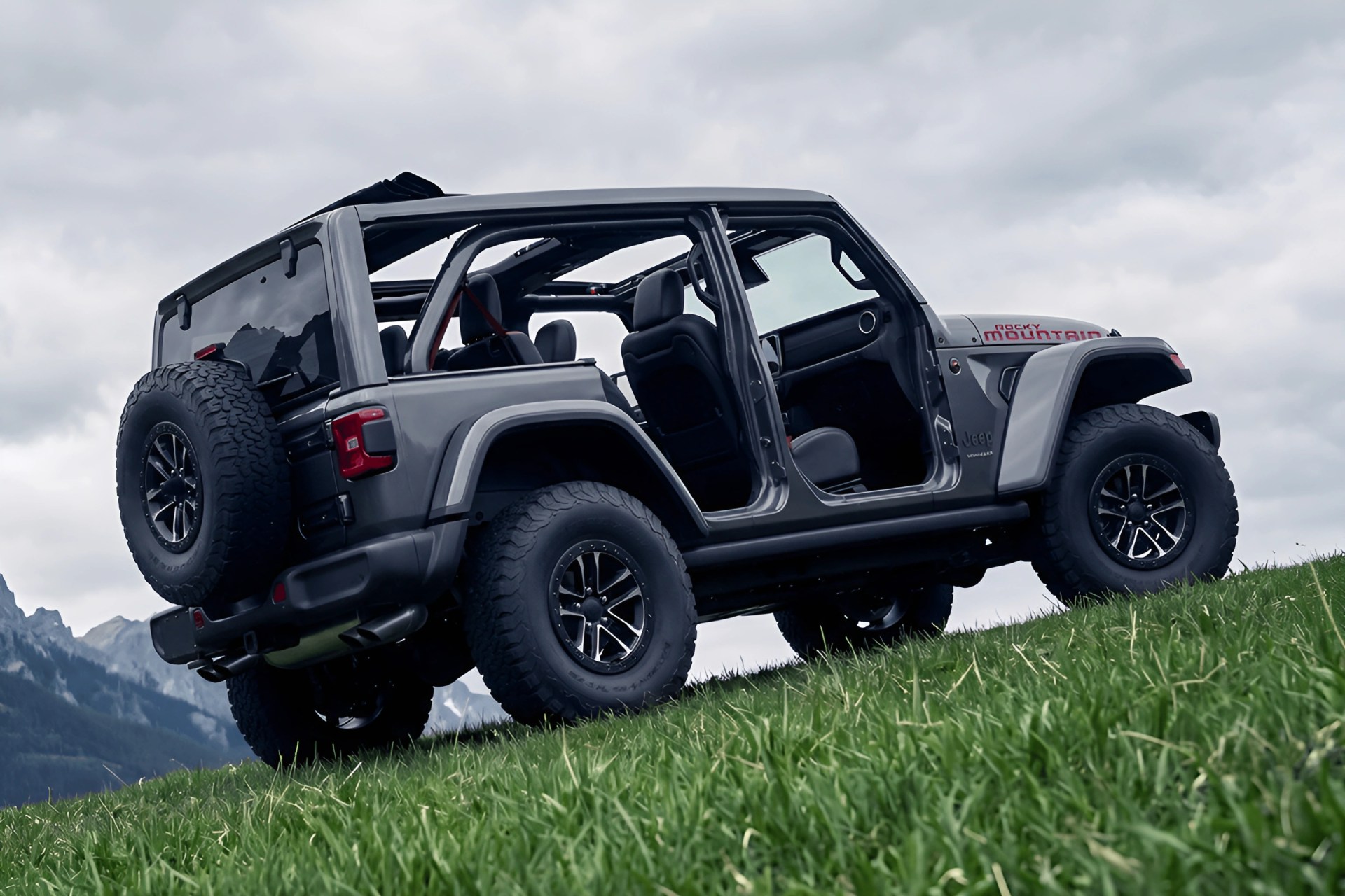 Gray Jeep Rocky Mountain edition with open doors and no roof on a grassy hill with mountains in the background.