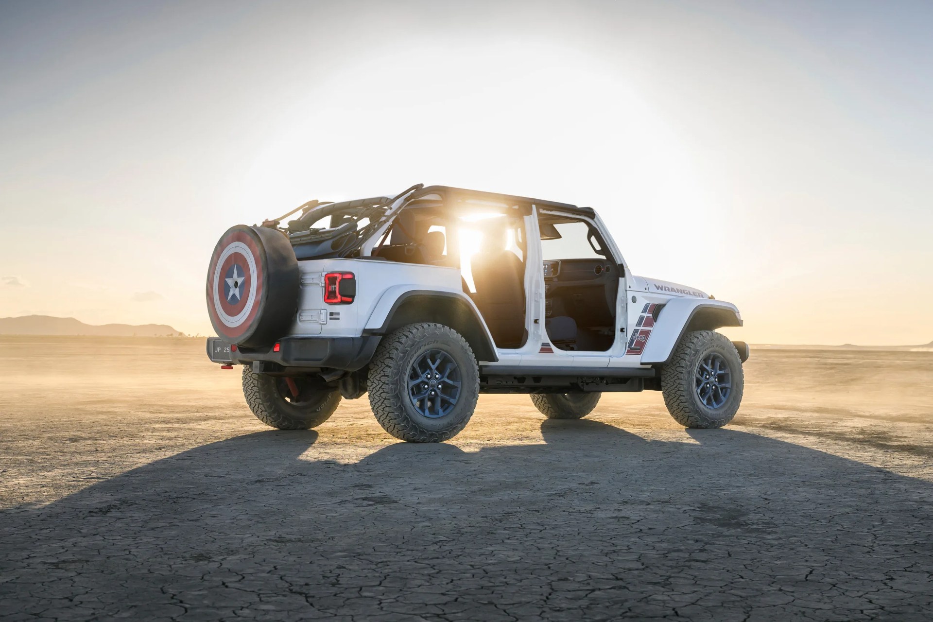 White Jeep Wrangler with open doors and Captain America shield tire cover on cracked desert ground at sunset.