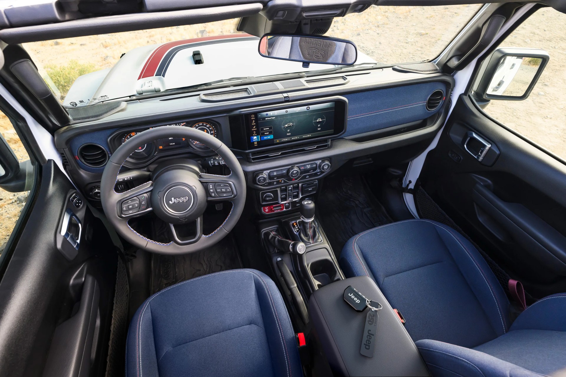 Interior of a Jeep vehicle showing a black steering wheel, digital dashboard, touchscreen display, blue fabric seats, and center console with Jeep key fob.