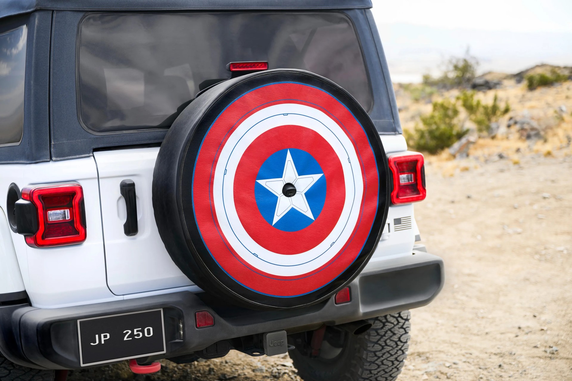 White Jeep with a Captain America shield-themed spare tire cover on a dirt road.