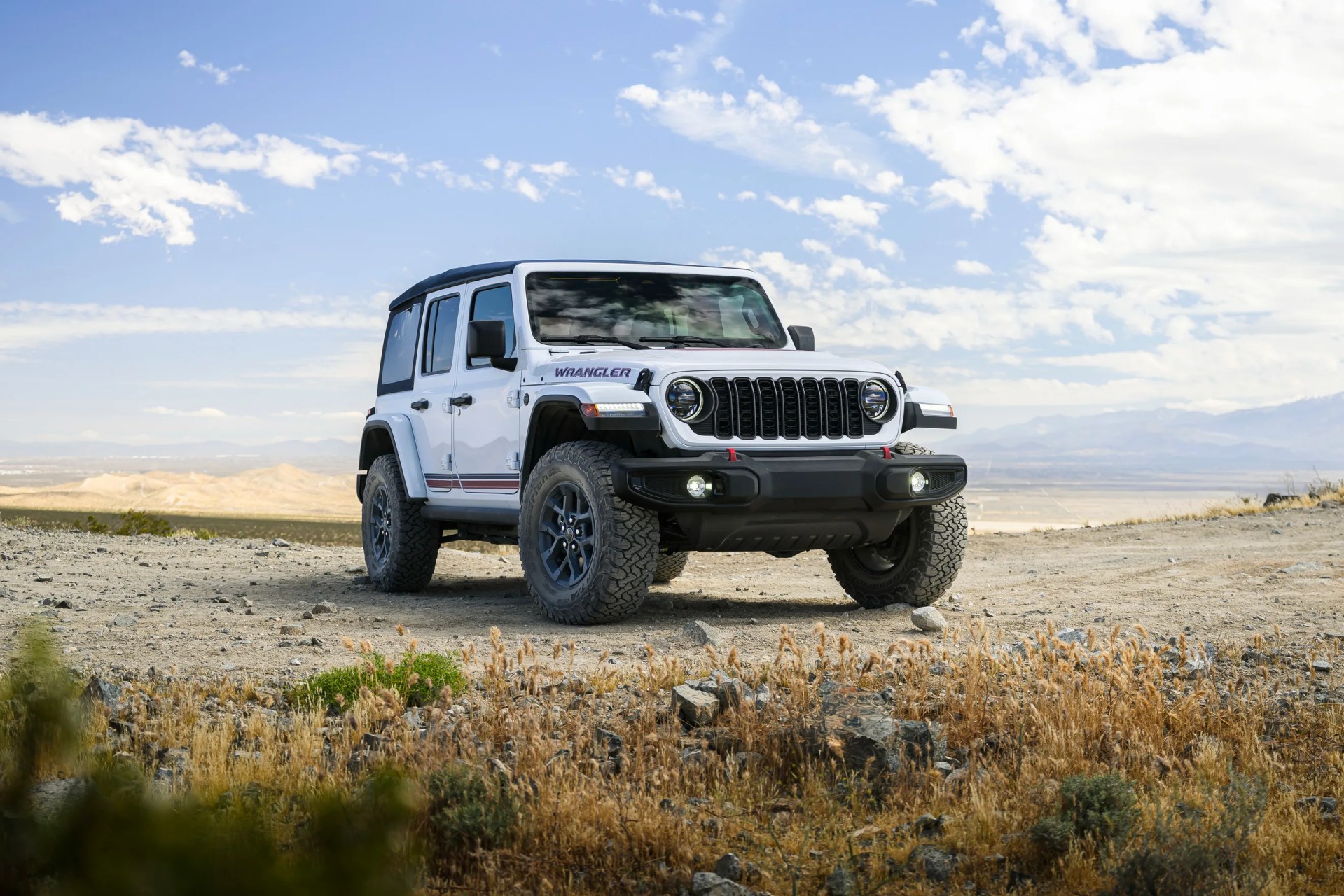 White Jeep Wrangler with black soft top parked on rocky terrain under a partly cloudy blue sky.