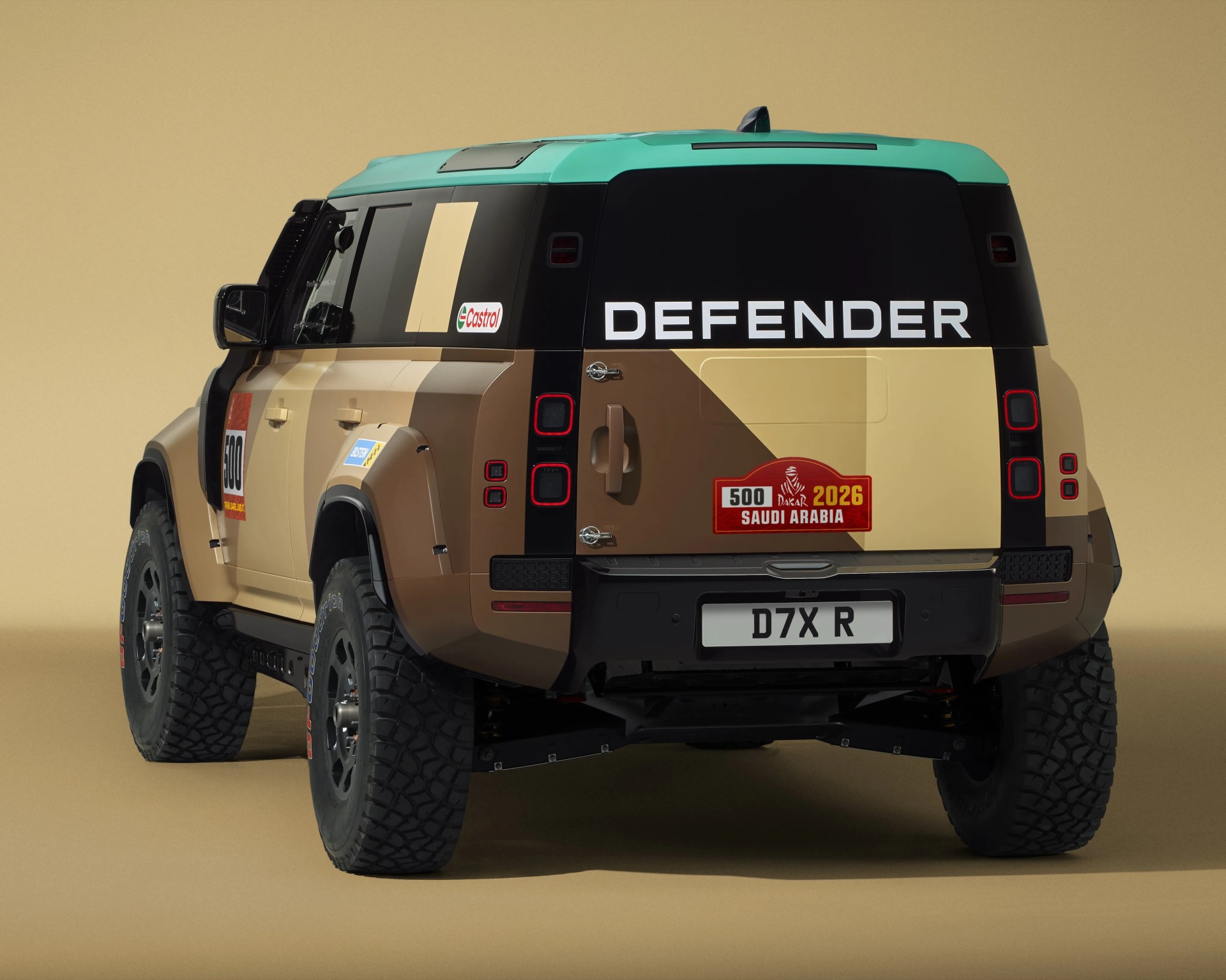 Rear view of a tan and brown Land Rover Defender with a turquoise roof, Dakar 2026 Saudi Arabia decal, and off-road tires.